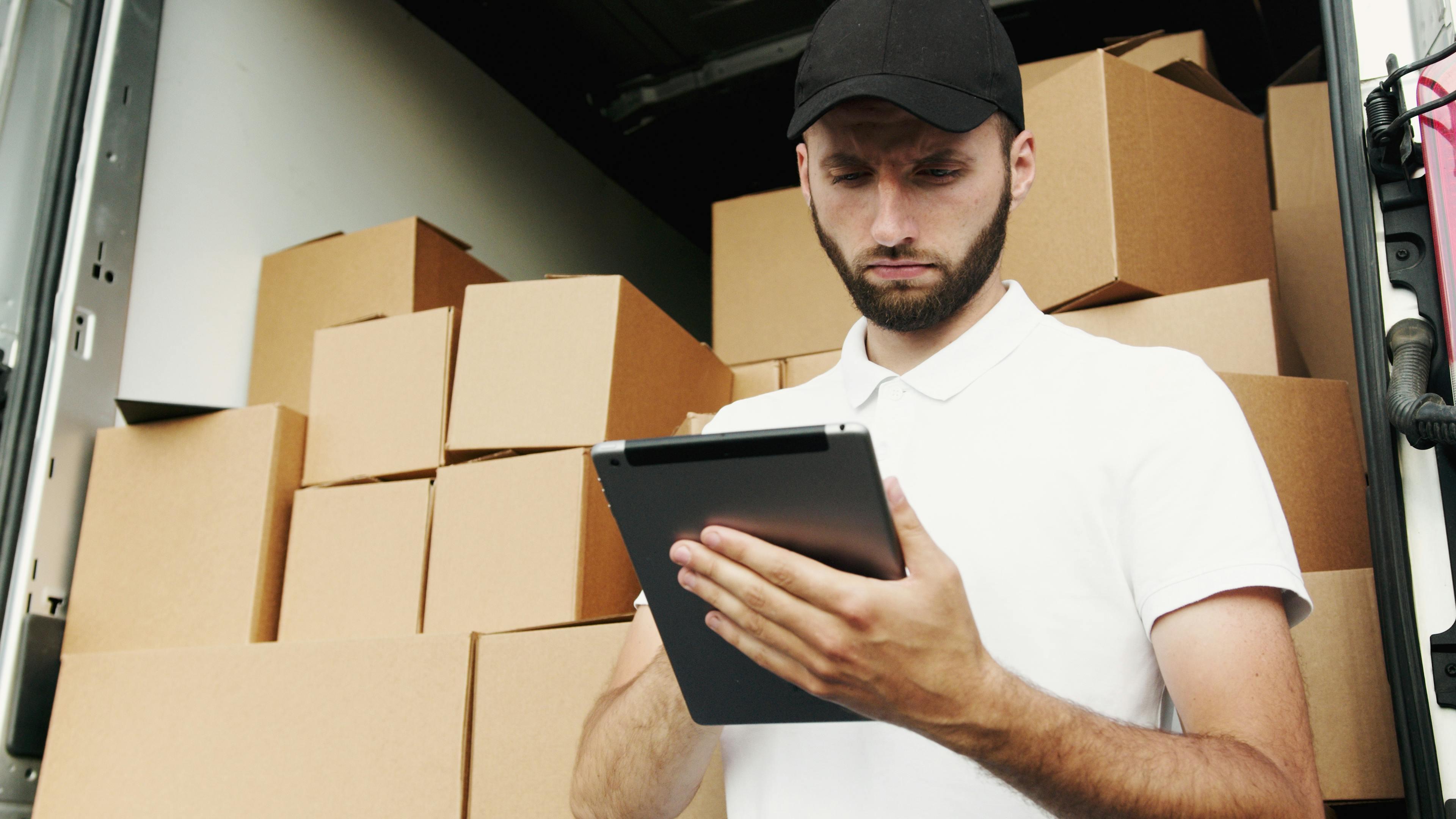 The man is checking his tablet in front of some packages.
