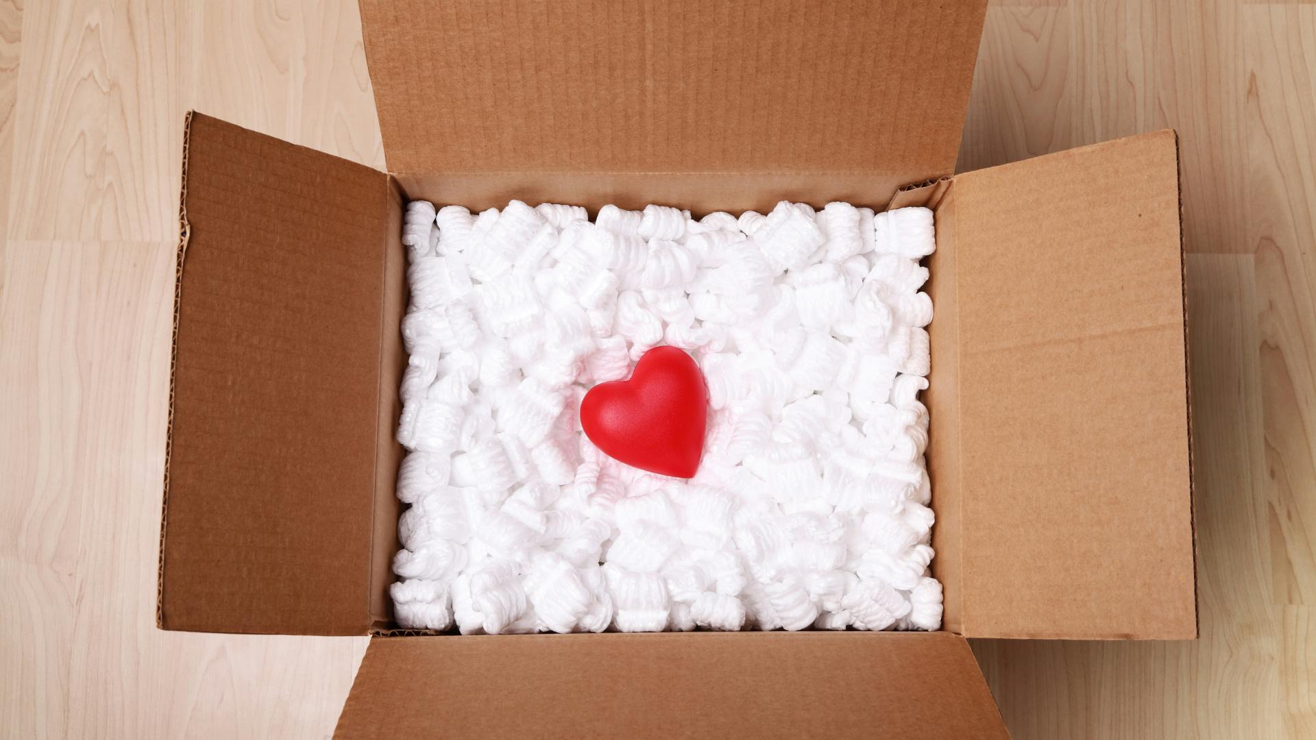 A shipping package filled with packing peanuts with a red heart on top of the packing peanuts. 