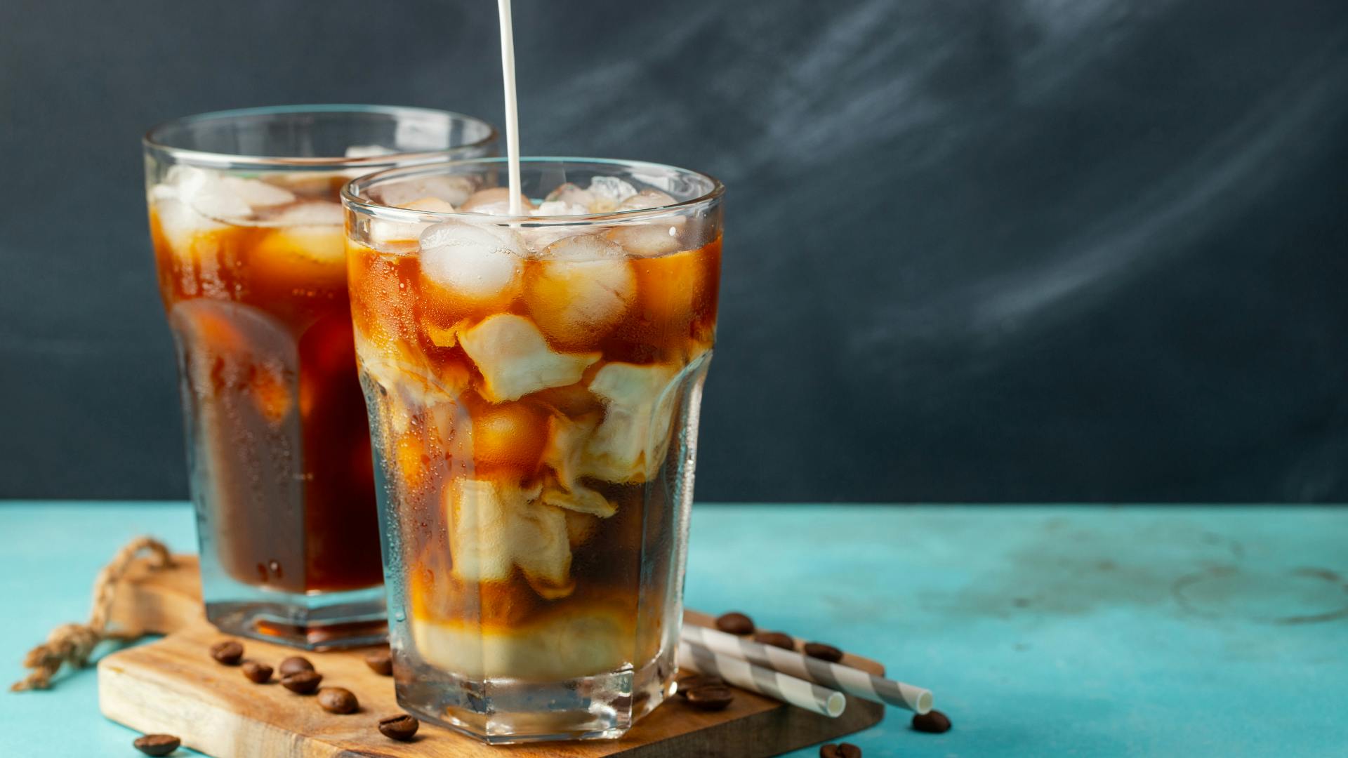 2 iced coffees with straw on a wooden plate with coffee grounds on the plate.