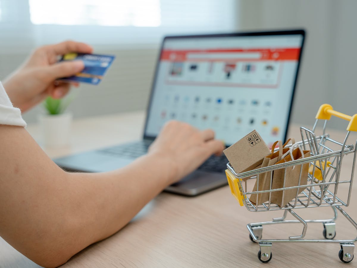 A person entering card details for online shopping on the background, and one miniature shopping cart with shopping bags. 