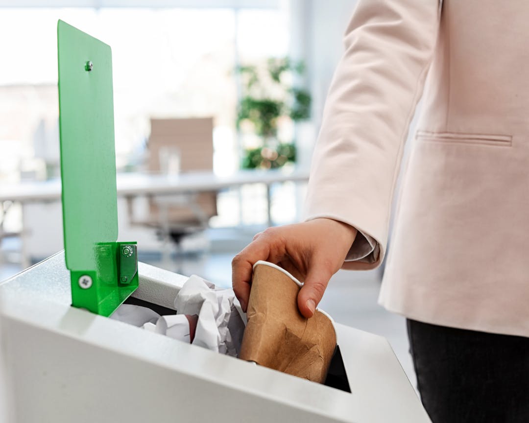 Person placing a disposable, single-use coffee cup into an open bin with a green lid.