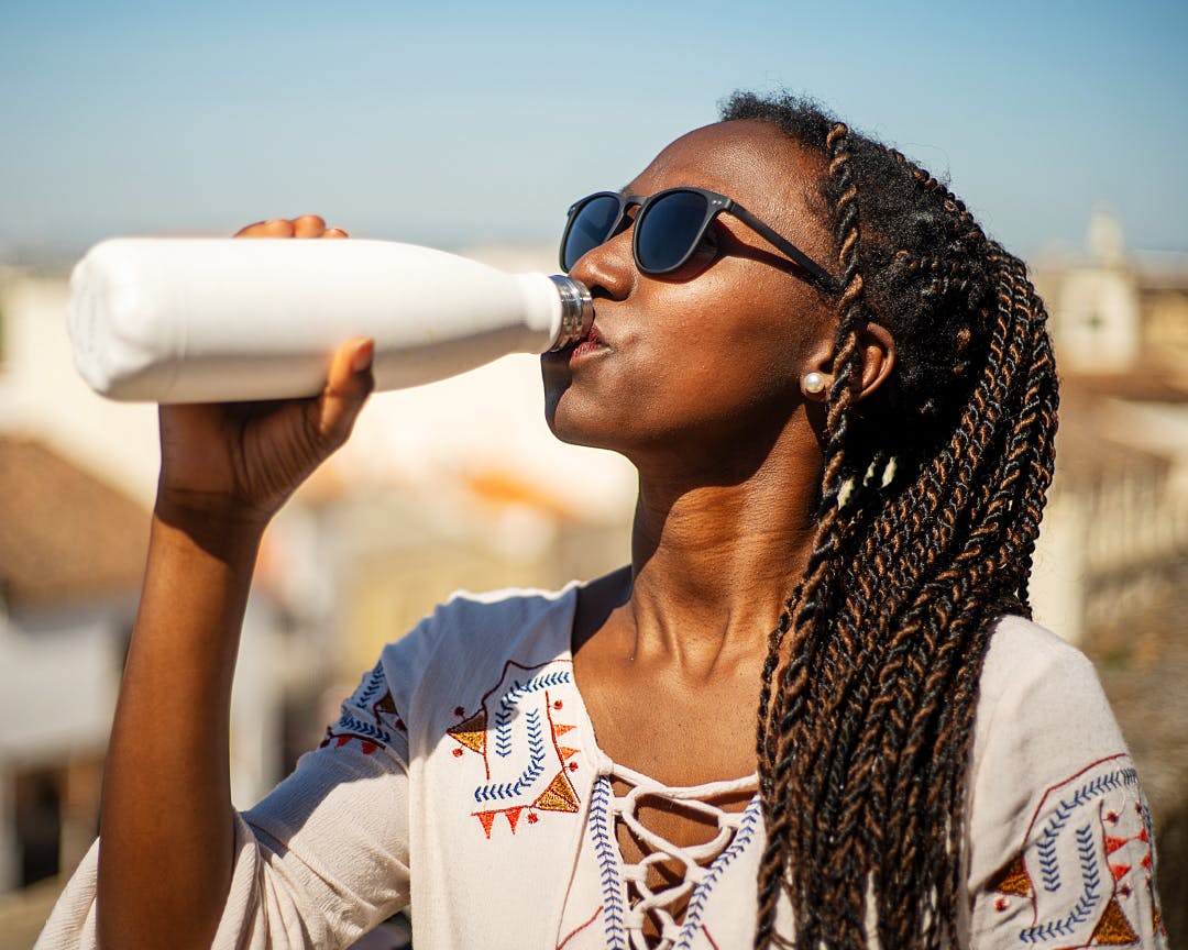 Person wearing sunglasses, drinking from a reusable bottle outdoors under a blue sky