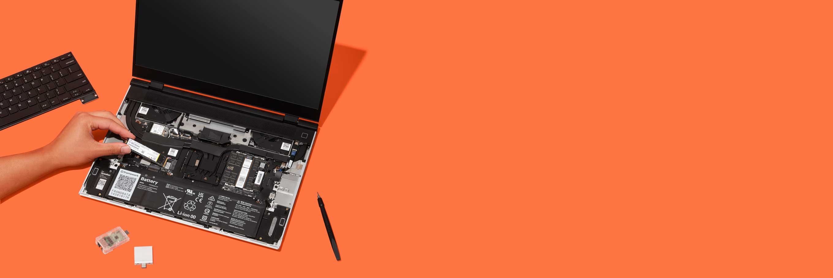 Top-down studio shot of a Framework Laptop 16 with a person installing a component on an orange backdrop.