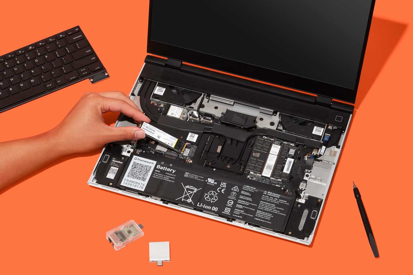 Top-down studio shot of a Framework Laptop 16 with a person installing a component on an orange backdrop.