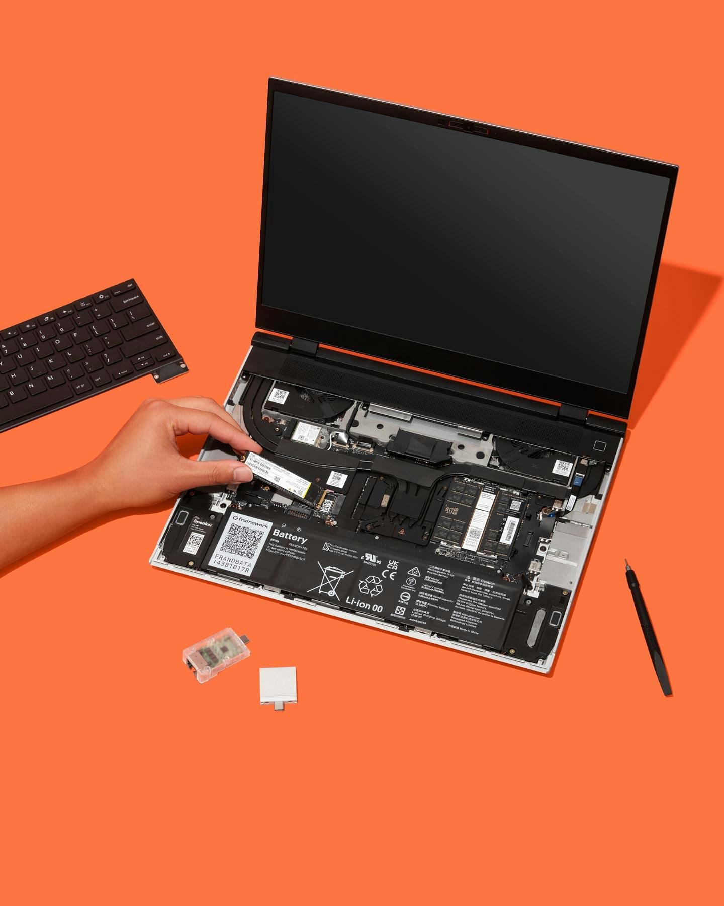 Top-down studio shot of a Framework Laptop 16 with a hand installing a component on an orange backdrop.