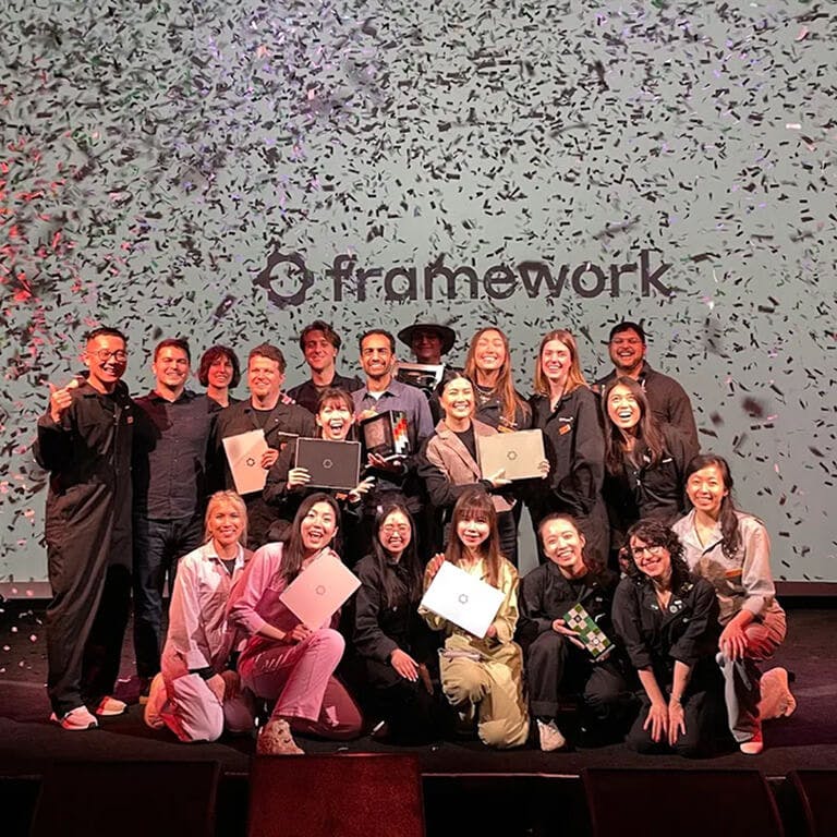 The Framework team standing on a stage with confetti raining down, holding Framework Laptops and awards in front of a large screen displaying the company logo.