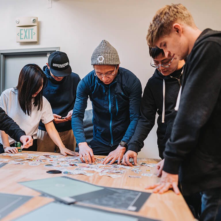 A group of employees gathered around a wooden table, looking down and sorting through a large pile of assorted stickers.