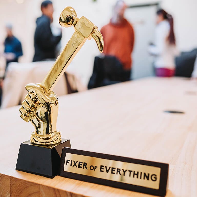 A close-up of a gold novelty trophy shaped like a fist holding a hammer, with a plaque at the base that reads "FIXER OF EVERYTHING."
