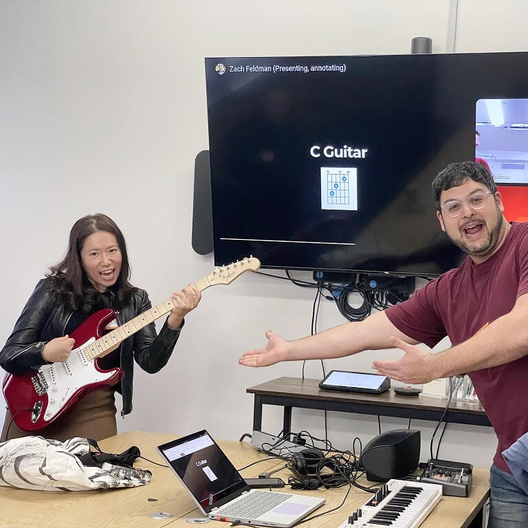 An employee holding a red electric guitar poses like a rockstar while a colleague gestures enthusiastically towards her.