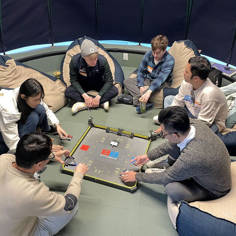 A group of Framework employees sitting in a circle, playing a tabletop game with miniature robots in a BattleBots arena.