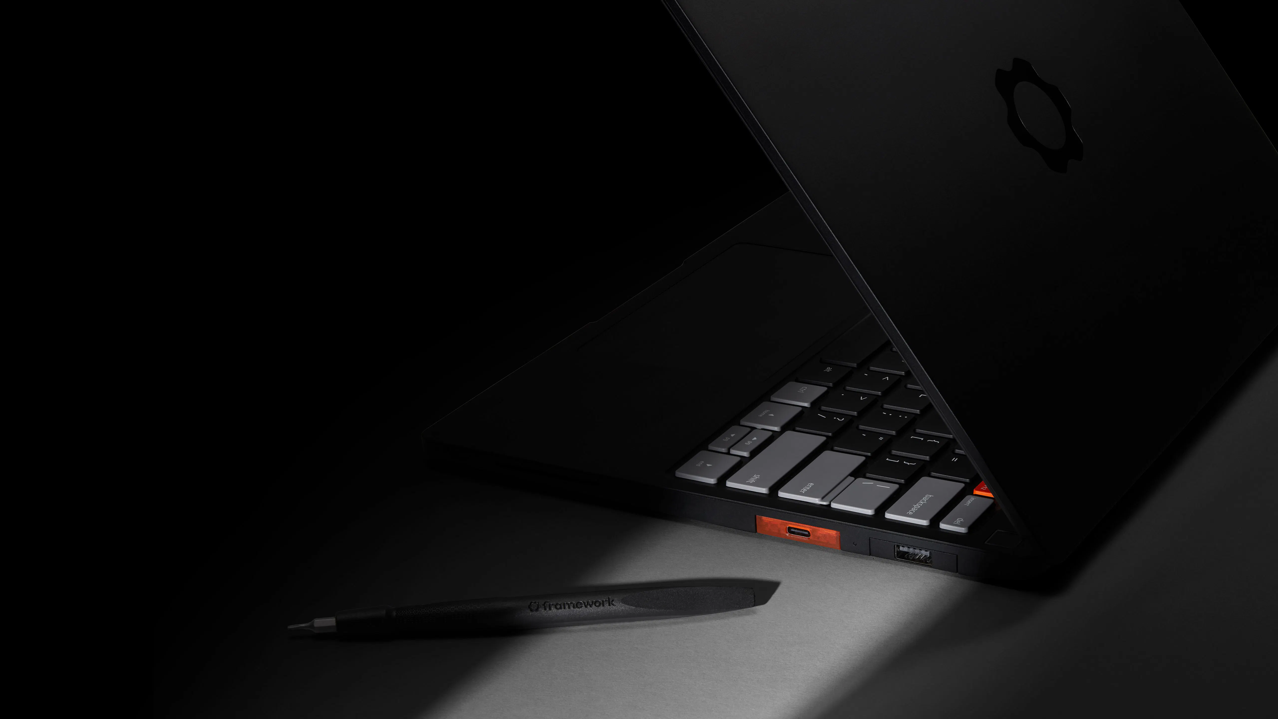  A high-angle, low-light shot of a sleek Framework Laptop 13 Pro partially open on a dark surface. A Framework screwdriver is placed closeby.