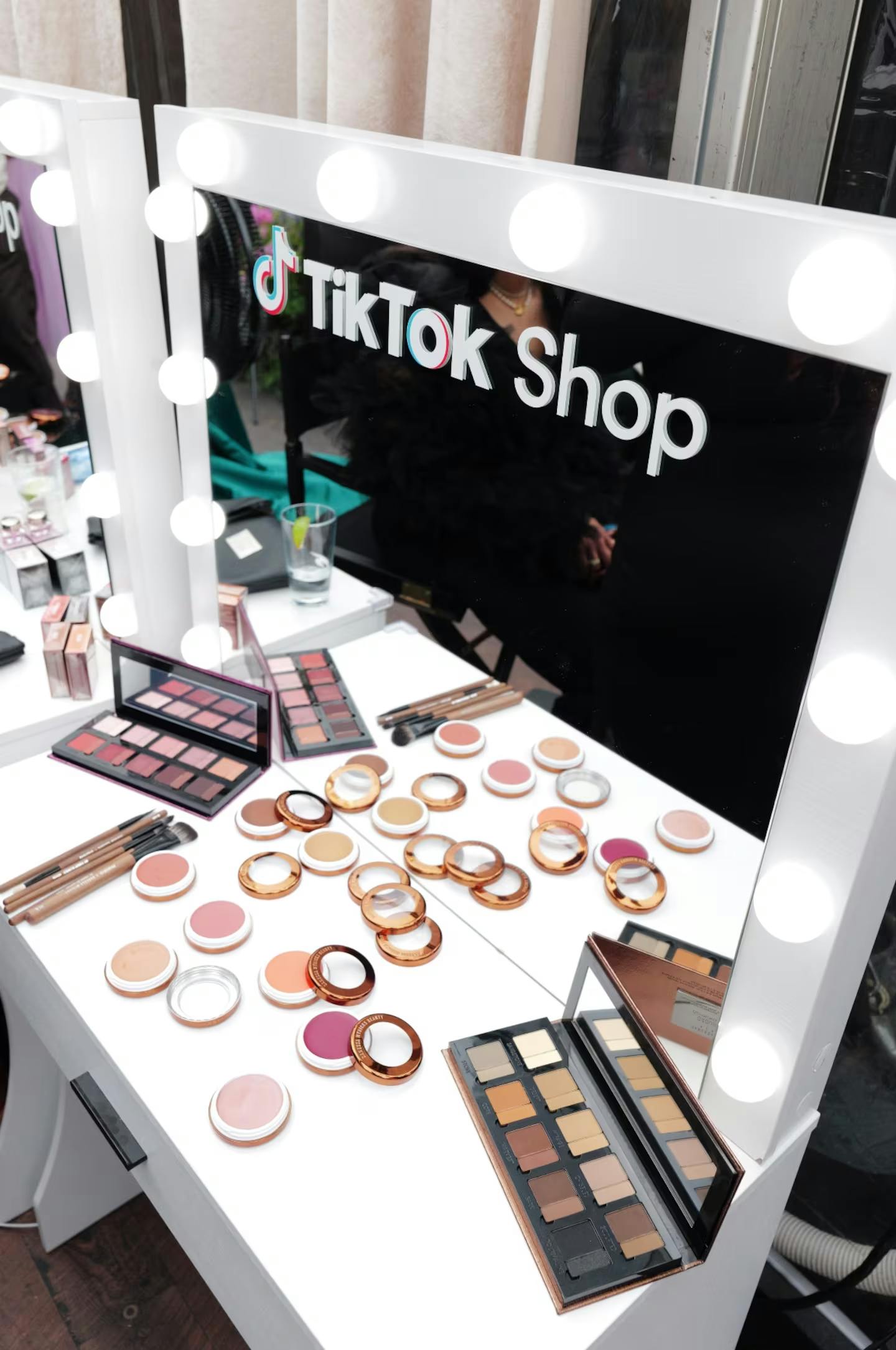 A white vanity table with a lighted mirror displaying the TikTok Shop logo. Cosmetics, including eyeshadow palettes and blushes, are arranged on the table.