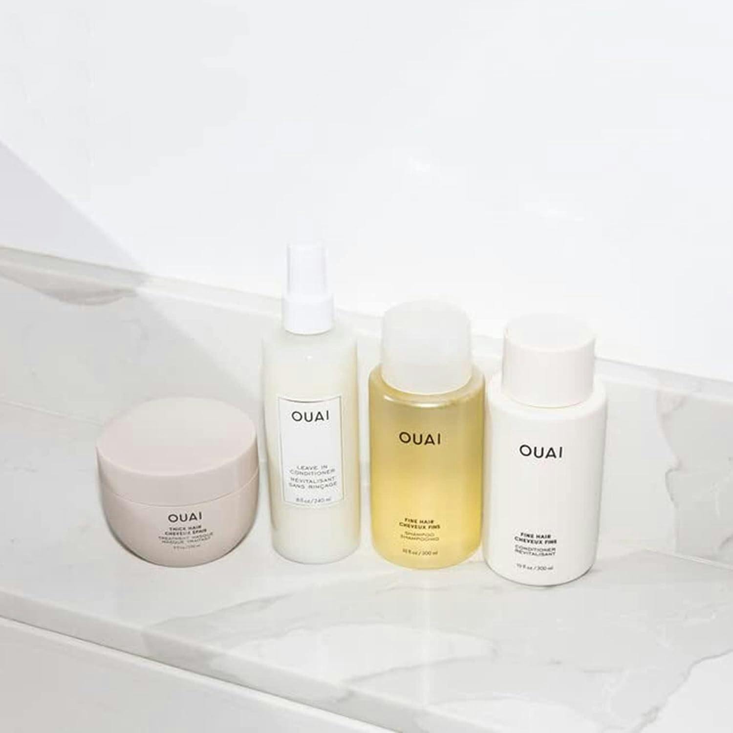 Four OUAI haircare products on a white marble table. From left to right: hair mask, leave in conditioner, fine hair shampoo and fine hair conditioner