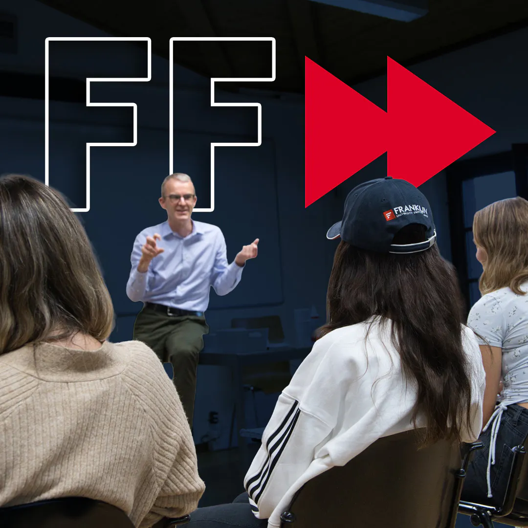 Presenter speaking to a seated audience in a modern lecture space, with bold “FF” graphics behind them.