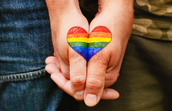 Two hands clasp, each partially painted with a joining side of a single heart that’s filled with a rainbow pattern.