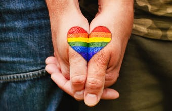 Two hands clasp, each partially painted with a joining side of a single heart that’s filled with a rainbow pattern.