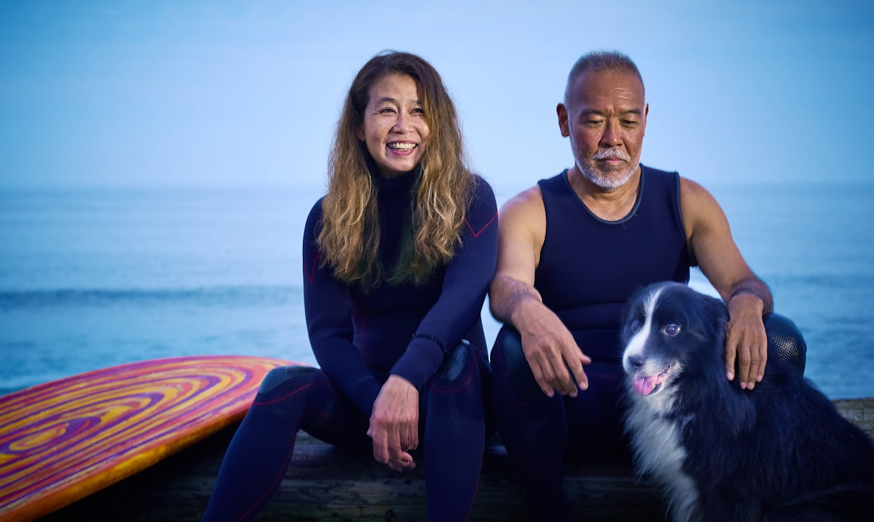 A surfer couple over 50 years old sits with their dog in front of the ocean.