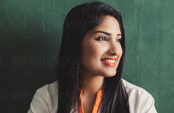 A young Indian woman wearing a colorful embroidered shirt sits in front of a green wall. 