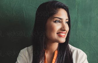 A young Indian woman wearing a colorful embroidered shirt sits in front of a green wall.