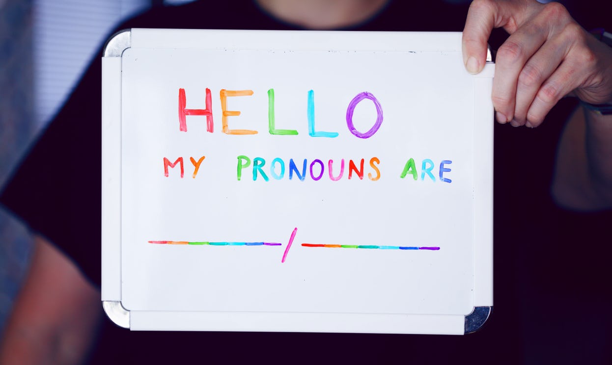 A person holding a whiteboard with “Hello, my pronouns are...” written on it in rainbow-colored ink.