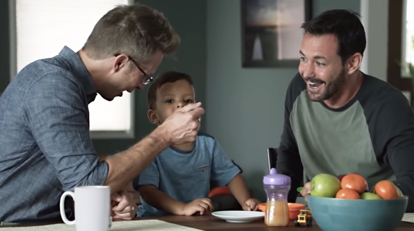 A gay couple enjoys feeding their child at the kitchen table.