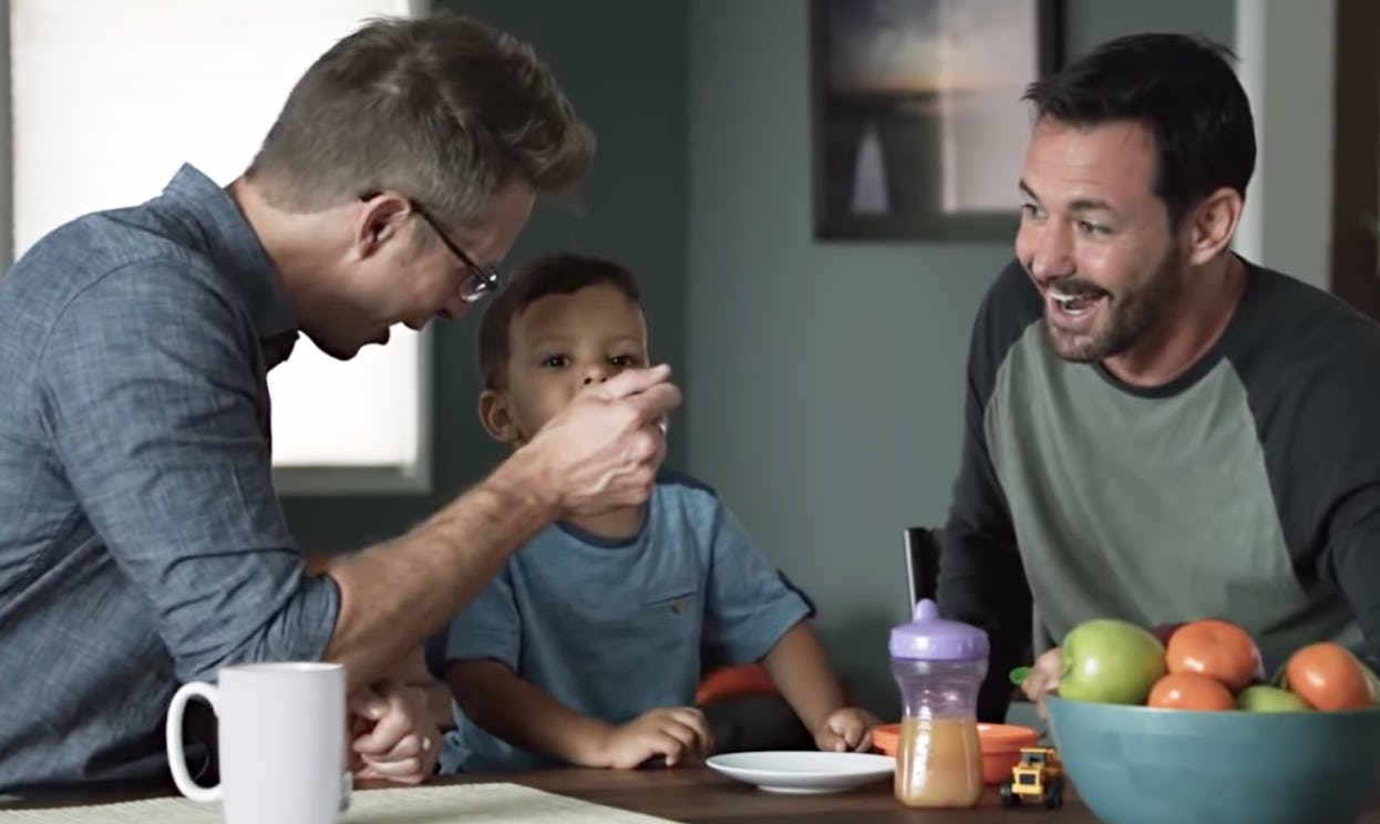 A gay couple enjoys feeding their child at the kitchen table.