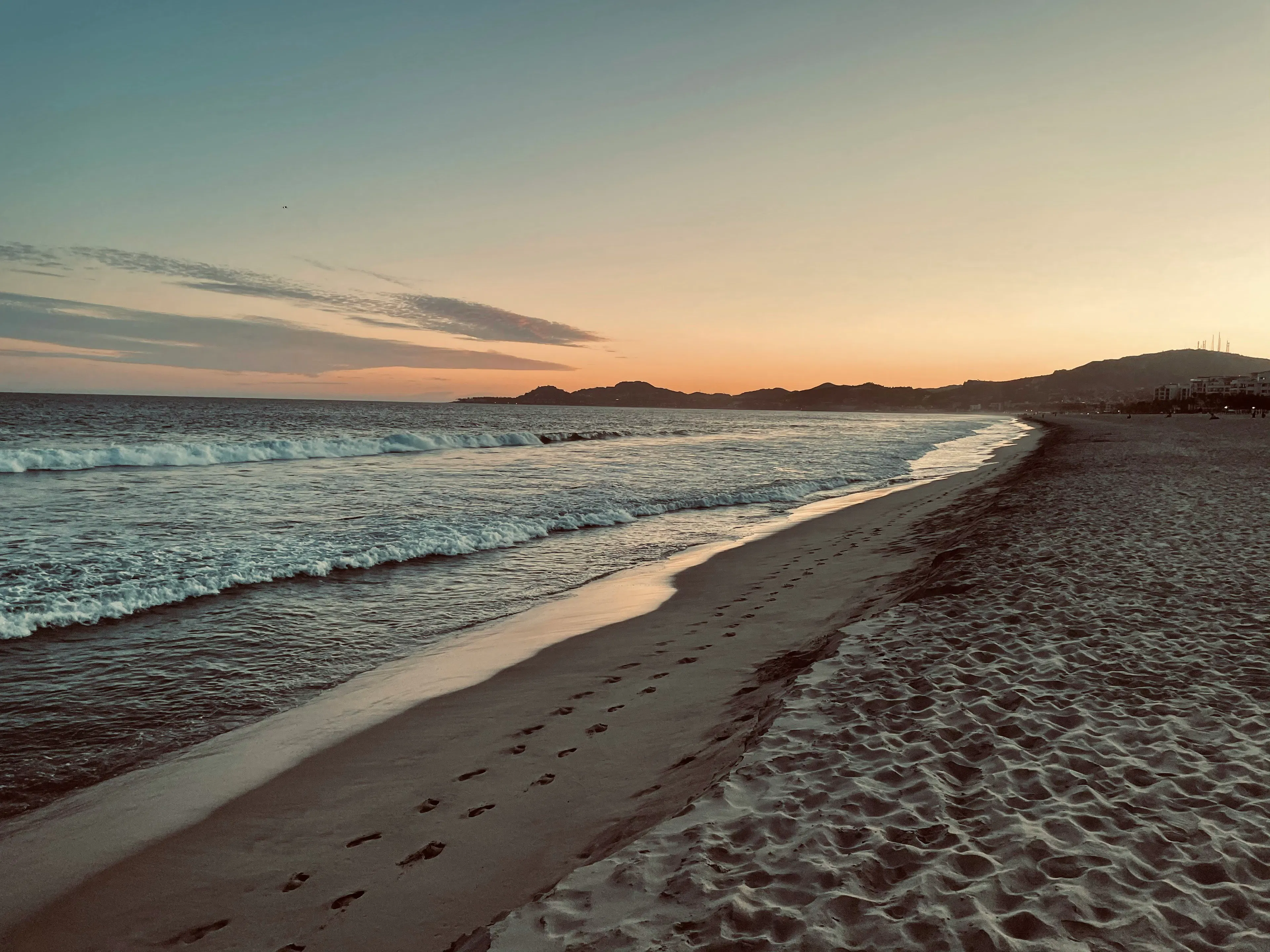 Ocean waves rolling onto a sandy beach at sunset with footprints along the shoreline and distant hills on the horizon.