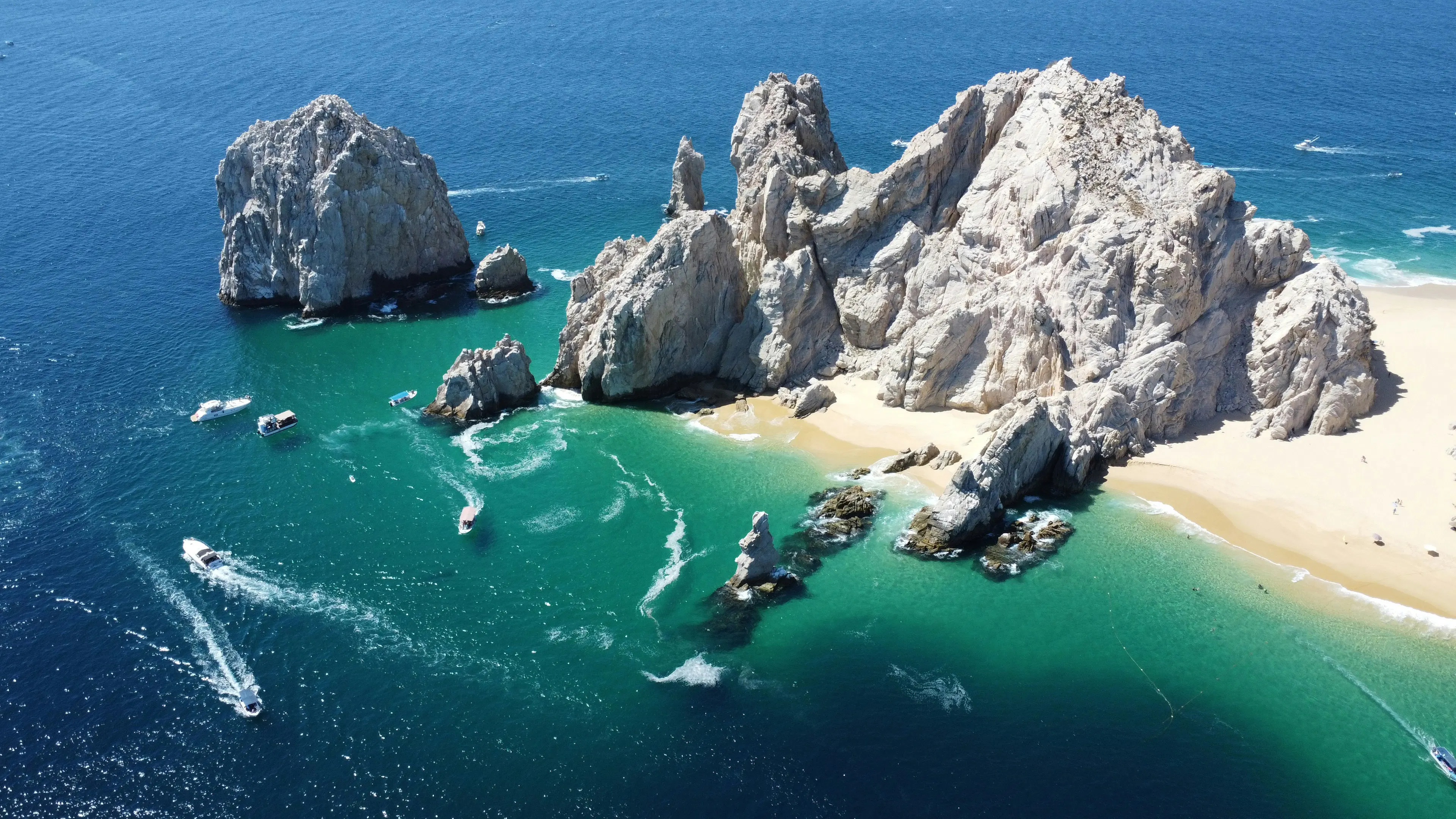 Aerial view of El Arco rock formations surrounded by turquoise water, sandy beaches, and boats near the shoreline.