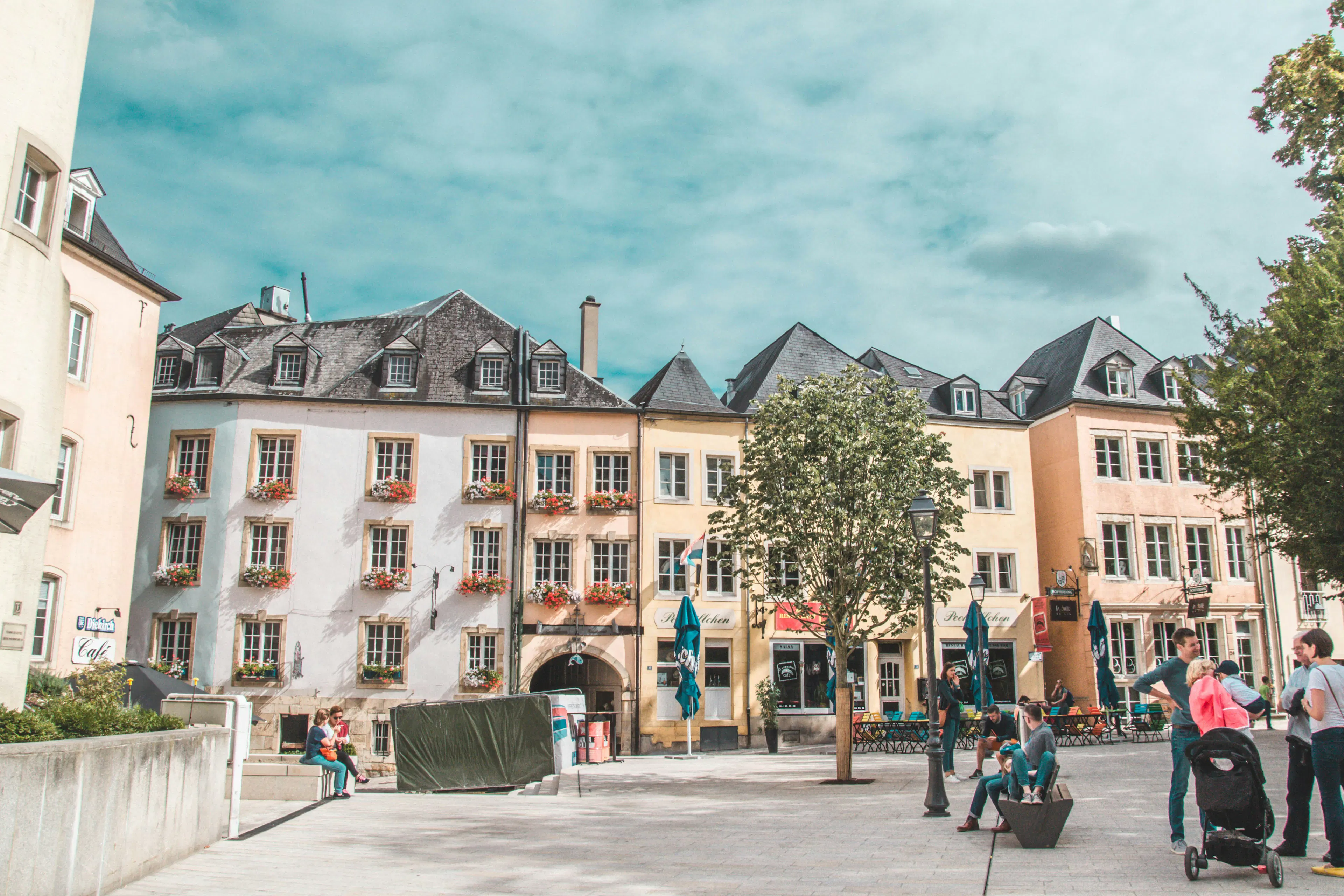Luxembourg city square with pastel buildings, cafe seating, trees, and people gathered near storefronts.