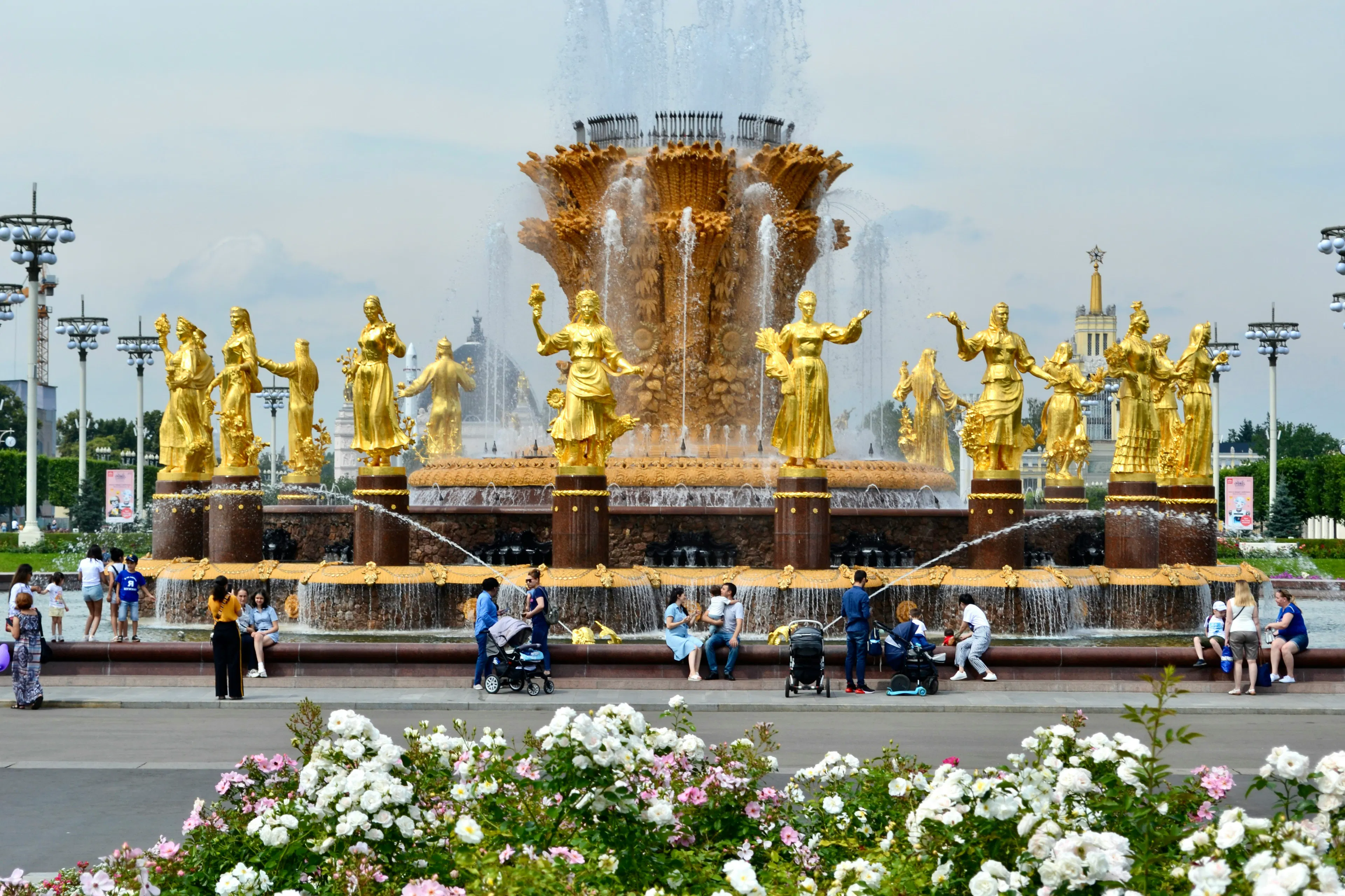 Large fountain in Moscow with gold statues, flowing water, flowers, and visitors gathered nearby.