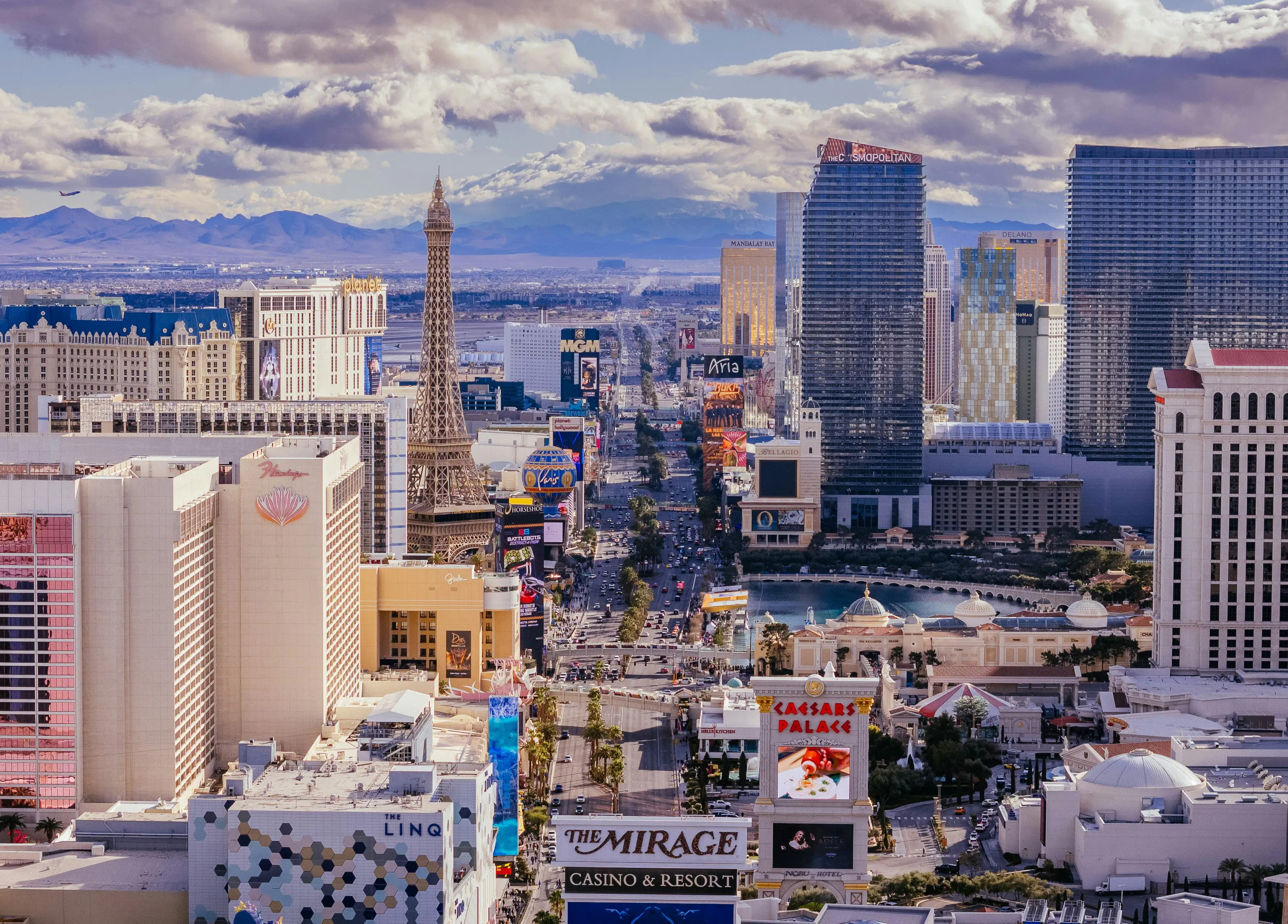 Aerial view of the Las Vegas Strip with resort towers, replica Eiffel Tower, and surrounding mountain ranges.