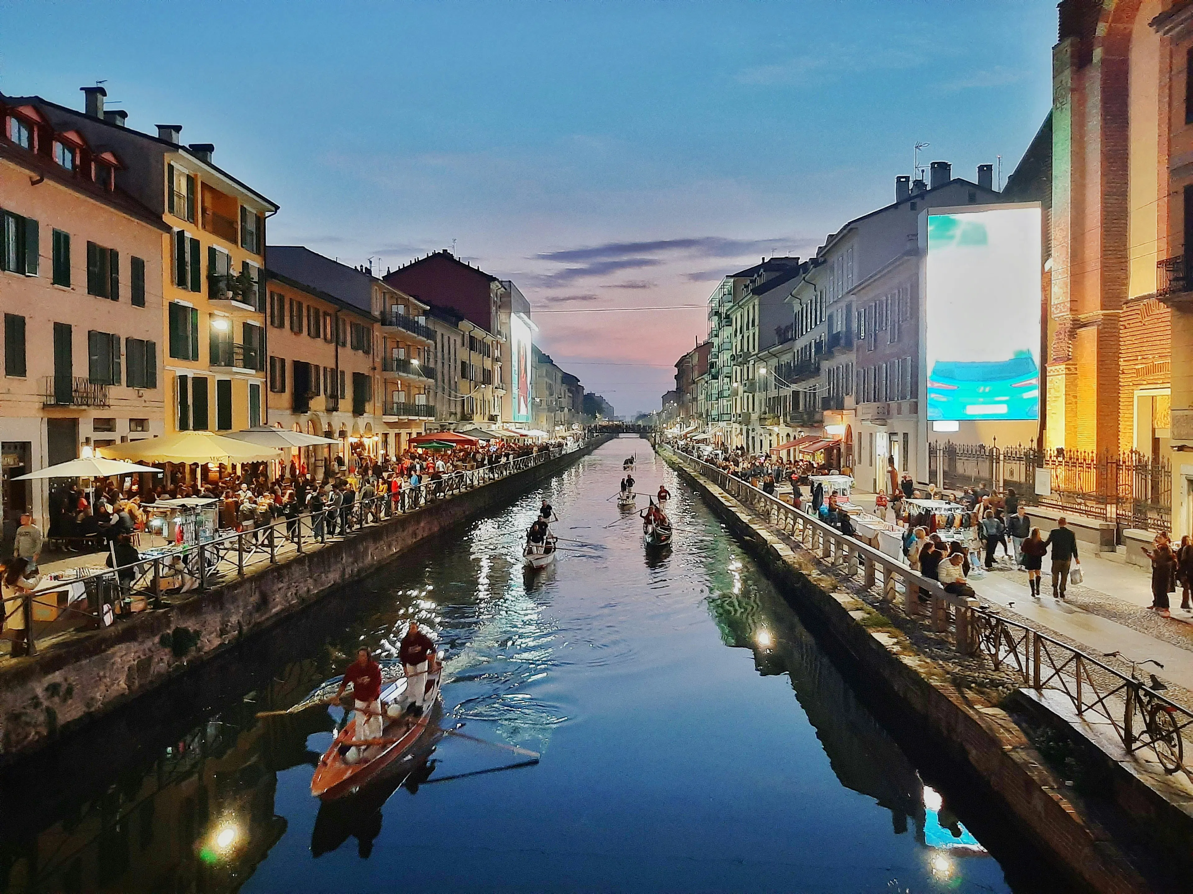 Canal in Milan’s Navigli district lined with restaurants and historic buildings, with small boats on the water and pedestrians along the waterfront at dusk.