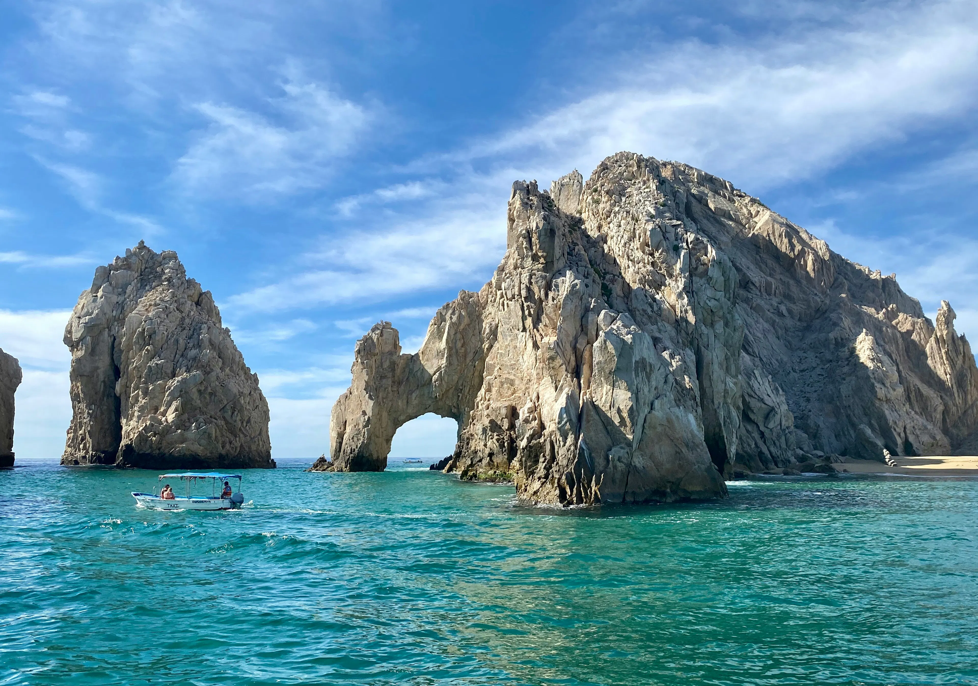 El Arco rock formation rising from the sea with turquoise water, coastal cliffs, and a small boat nearby.