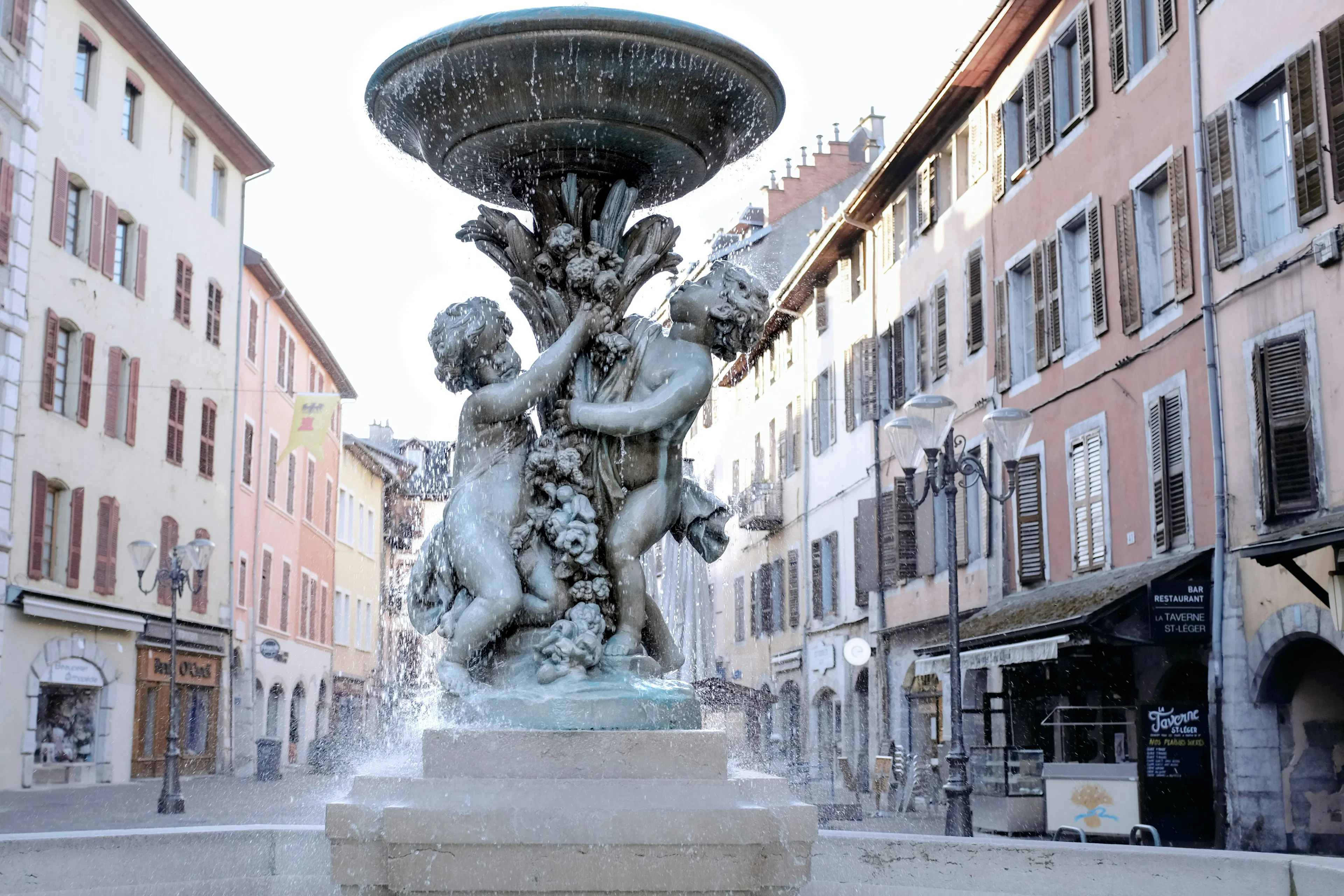 Stone fountain with sculpted figures in a city square lined with pastel buildings and shuttered windows.