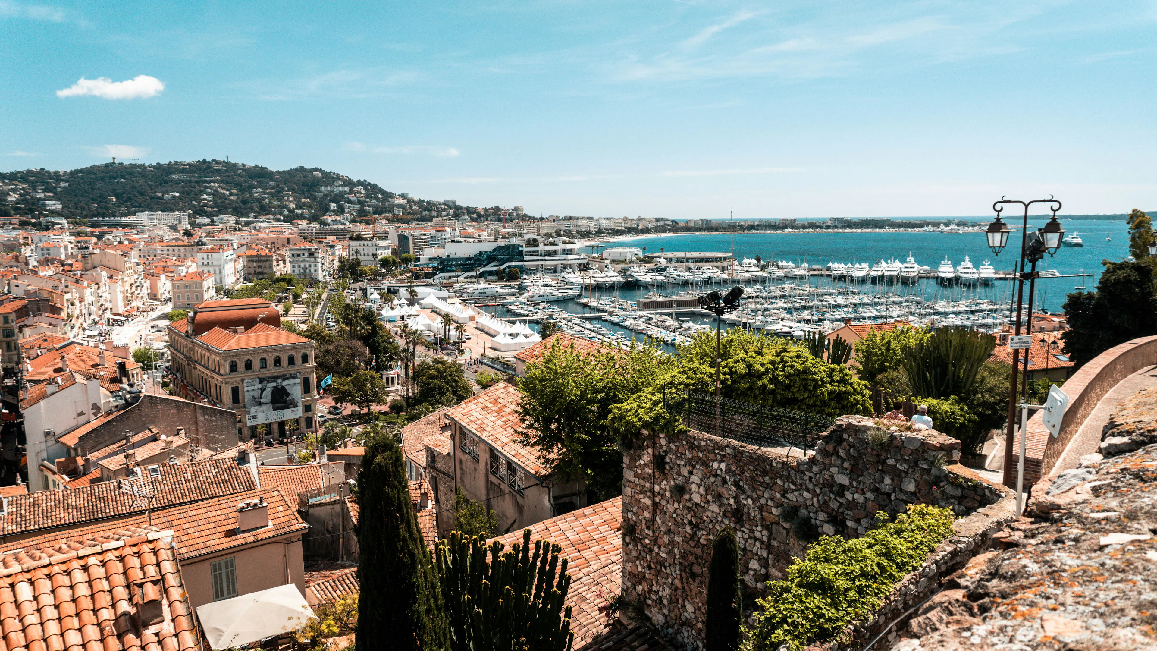 Aerial view of Cannes with red-roofed buildings, a marina, blue coastline, and hillside homes.