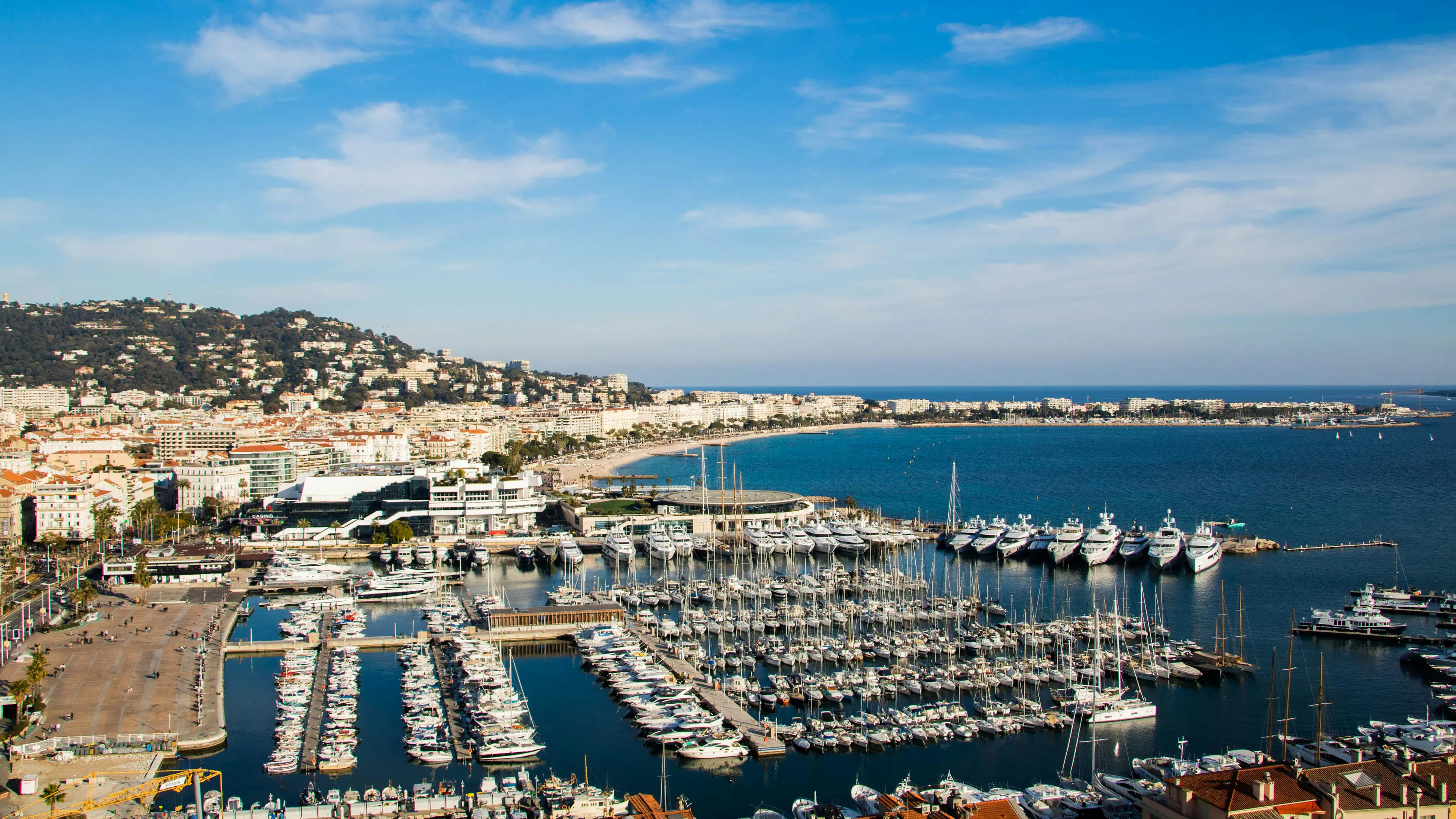Cannes harbor with yachts, sailboats, beachfront buildings, hills, and blue sea under a clear sky.