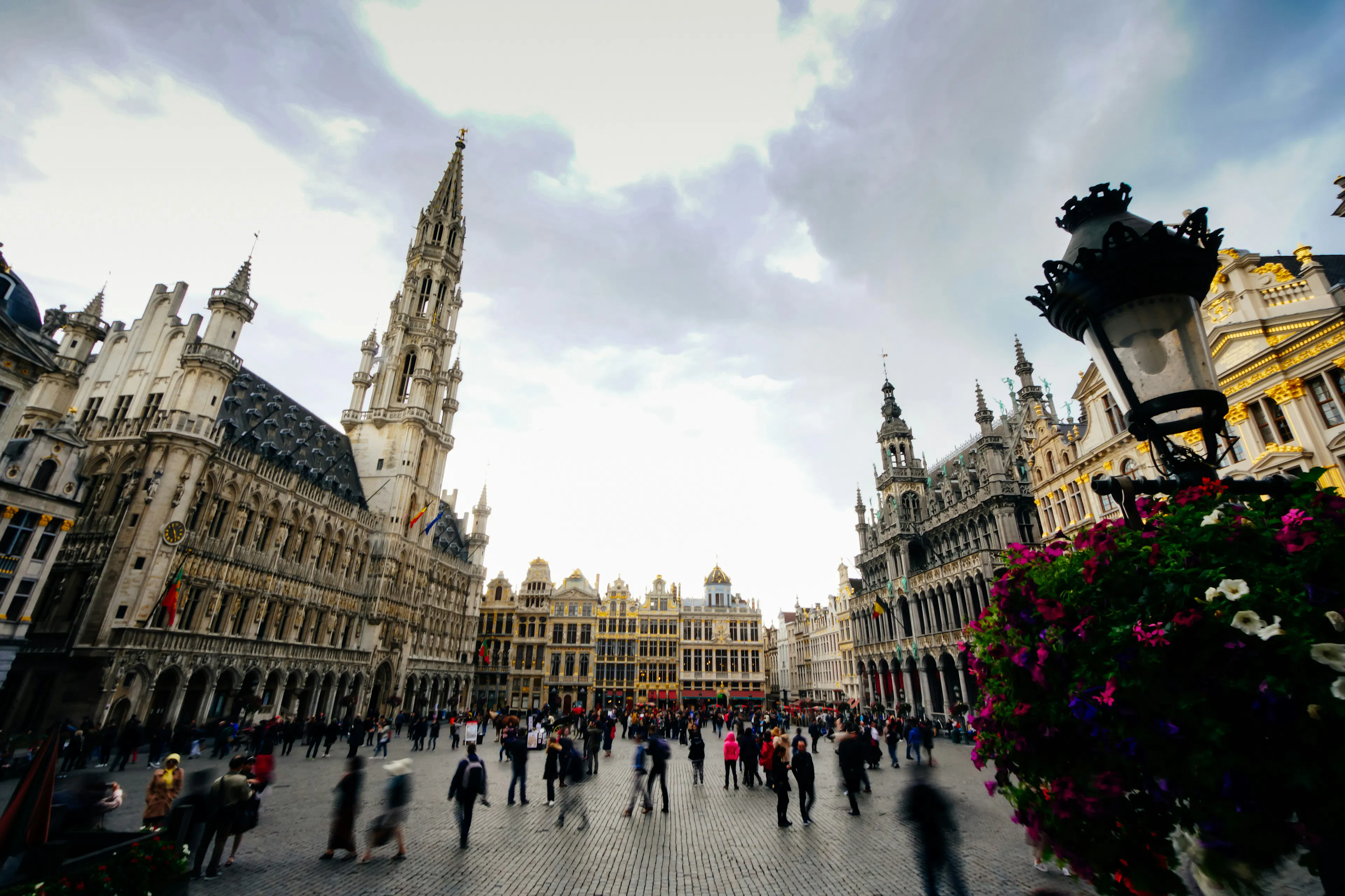Busy Brussels square with historic buildings, a tall tower, pedestrians, flowers, and a cloudy sky.