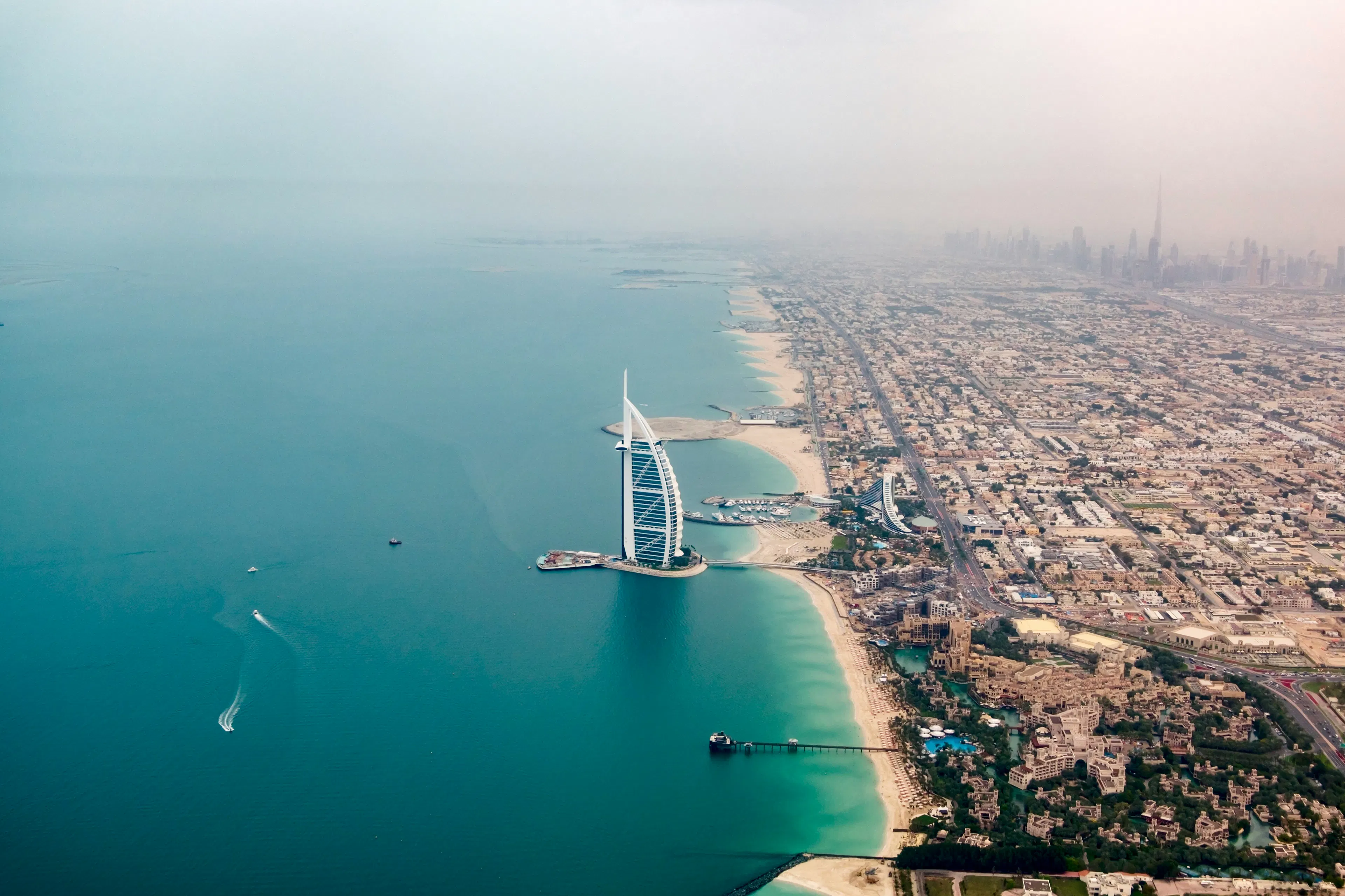 Aerial view of the Burj Al Arab hotel along Dubai’s coastline with turquoise water and city skyline in the distance.