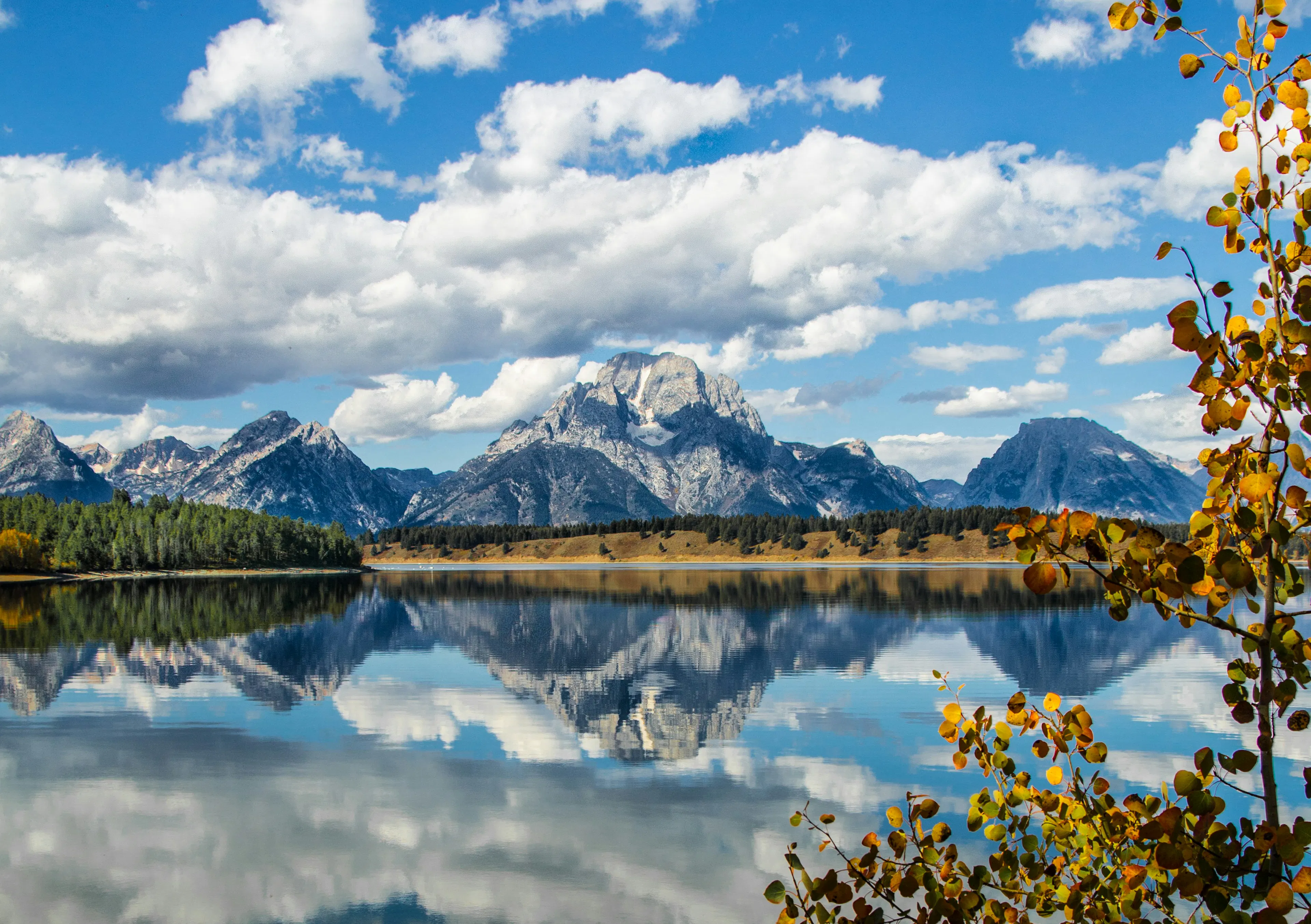 Snow-dusted mountain range reflected in a calm lake beneath scattered clouds and autumn trees.