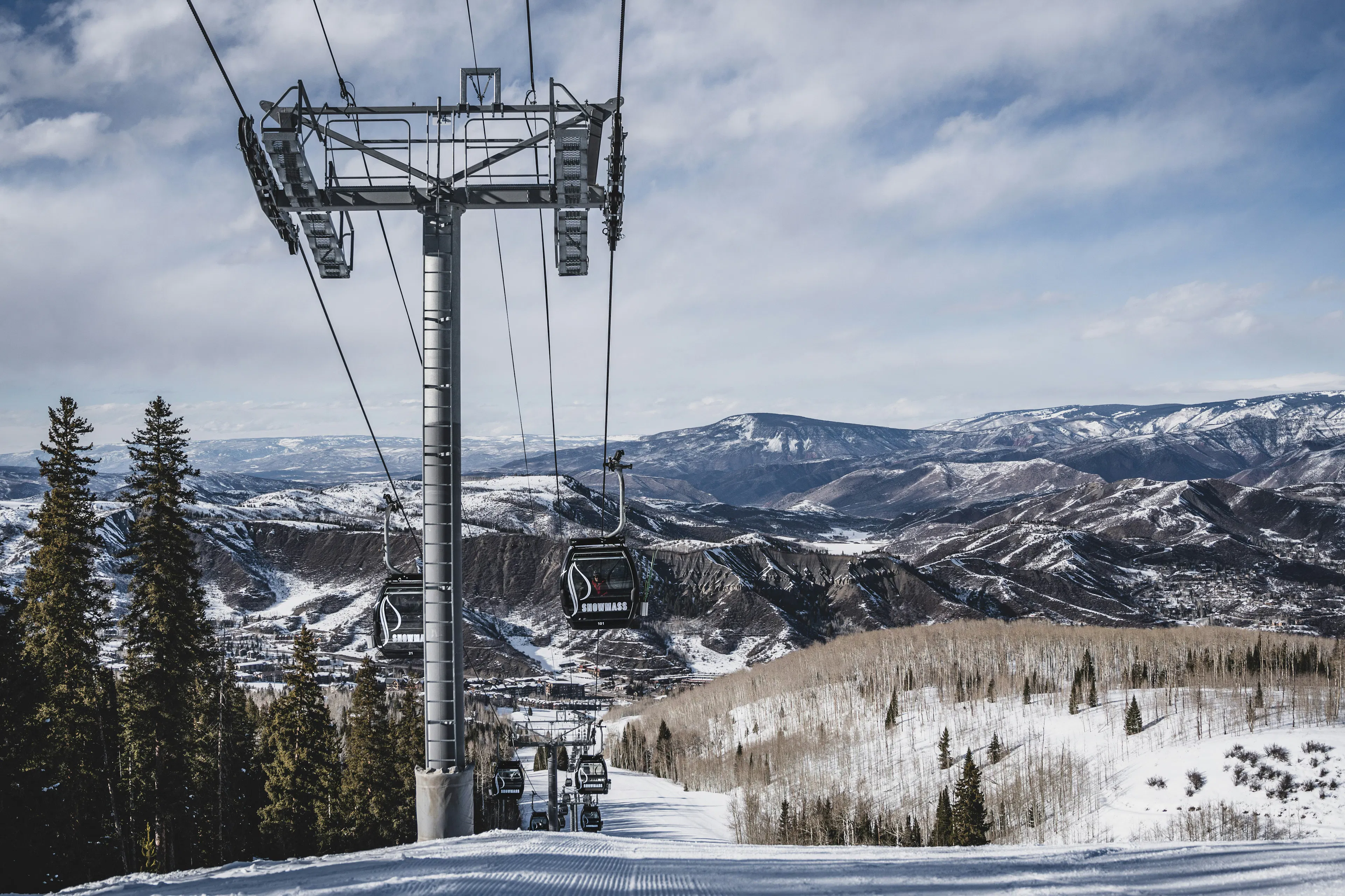 Gondola lift above a snow-covered ski slope with mountain ridges and evergreen trees surrounding Aspen.