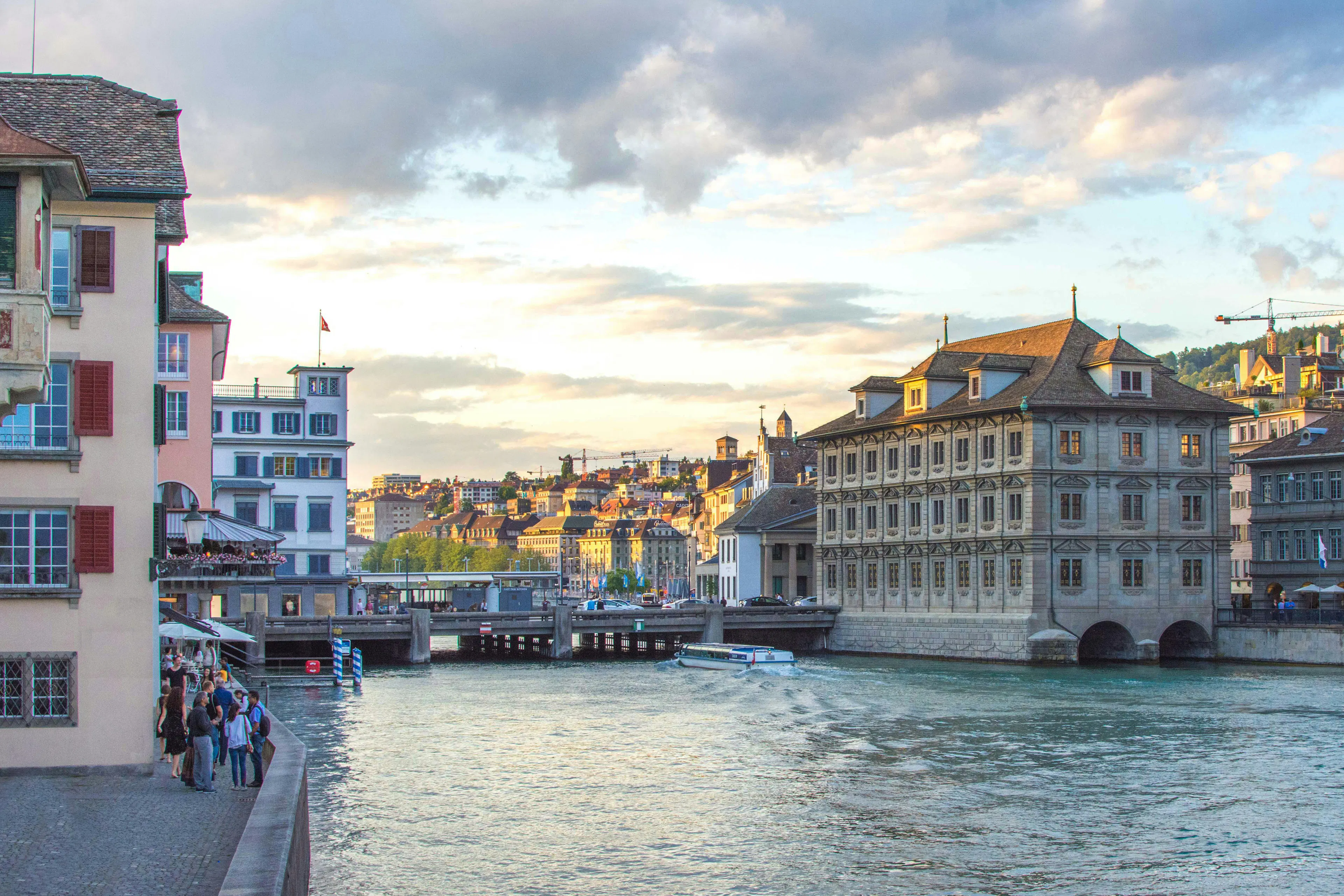 Historic buildings line the Limmat River in Zurich, Switzerland, as a boat travels along the water beneath a cloudy evening sky.