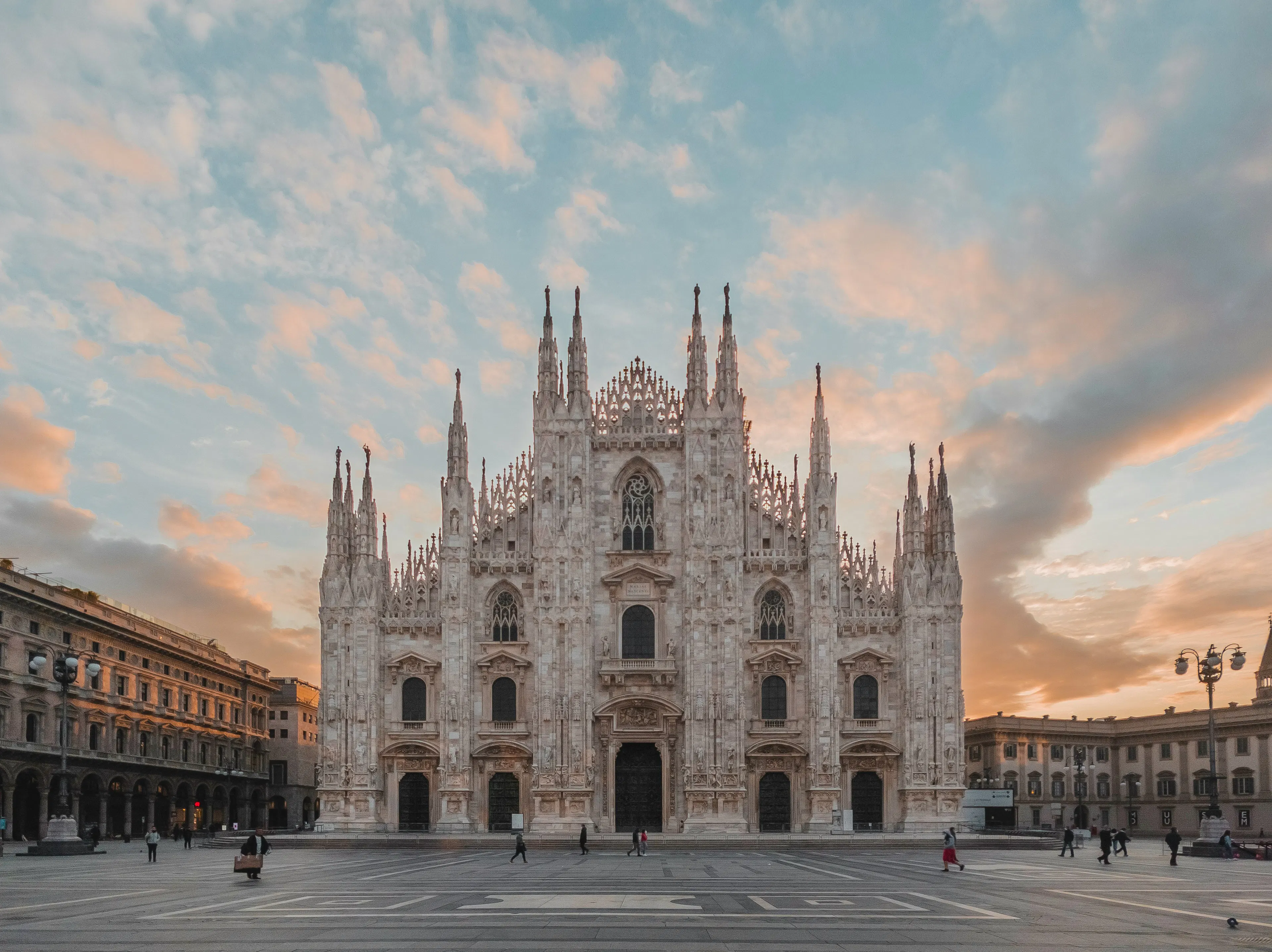 Milan Cathedral (Duomo di Milano) in Piazza del Duomo, featuring ornate Gothic architecture with people walking across the public square at sunset.