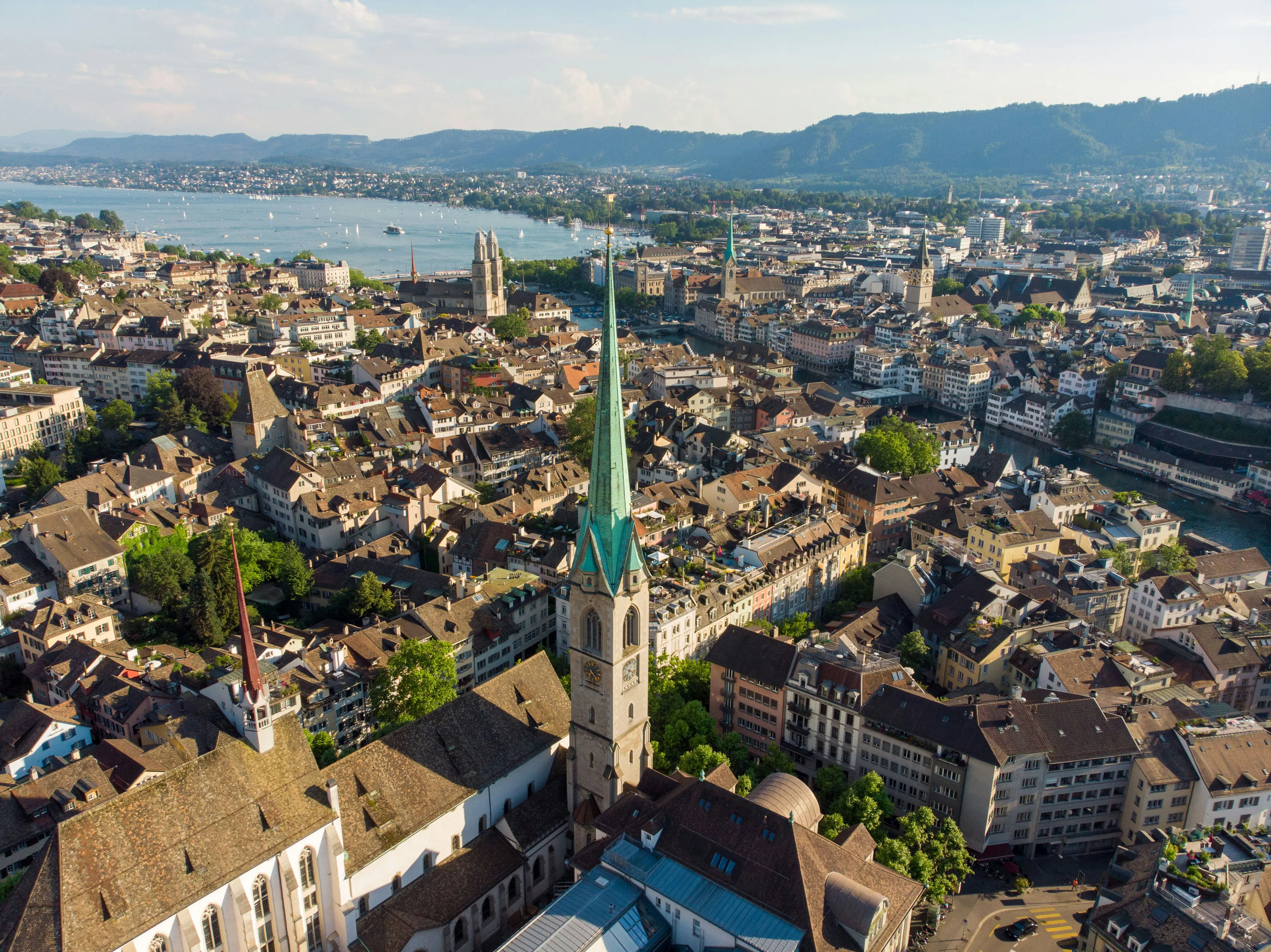 Aerial view of Zurich, Switzerland, featuring historic buildings, church spires, and Lake Zurich extending toward forested hills in the background.