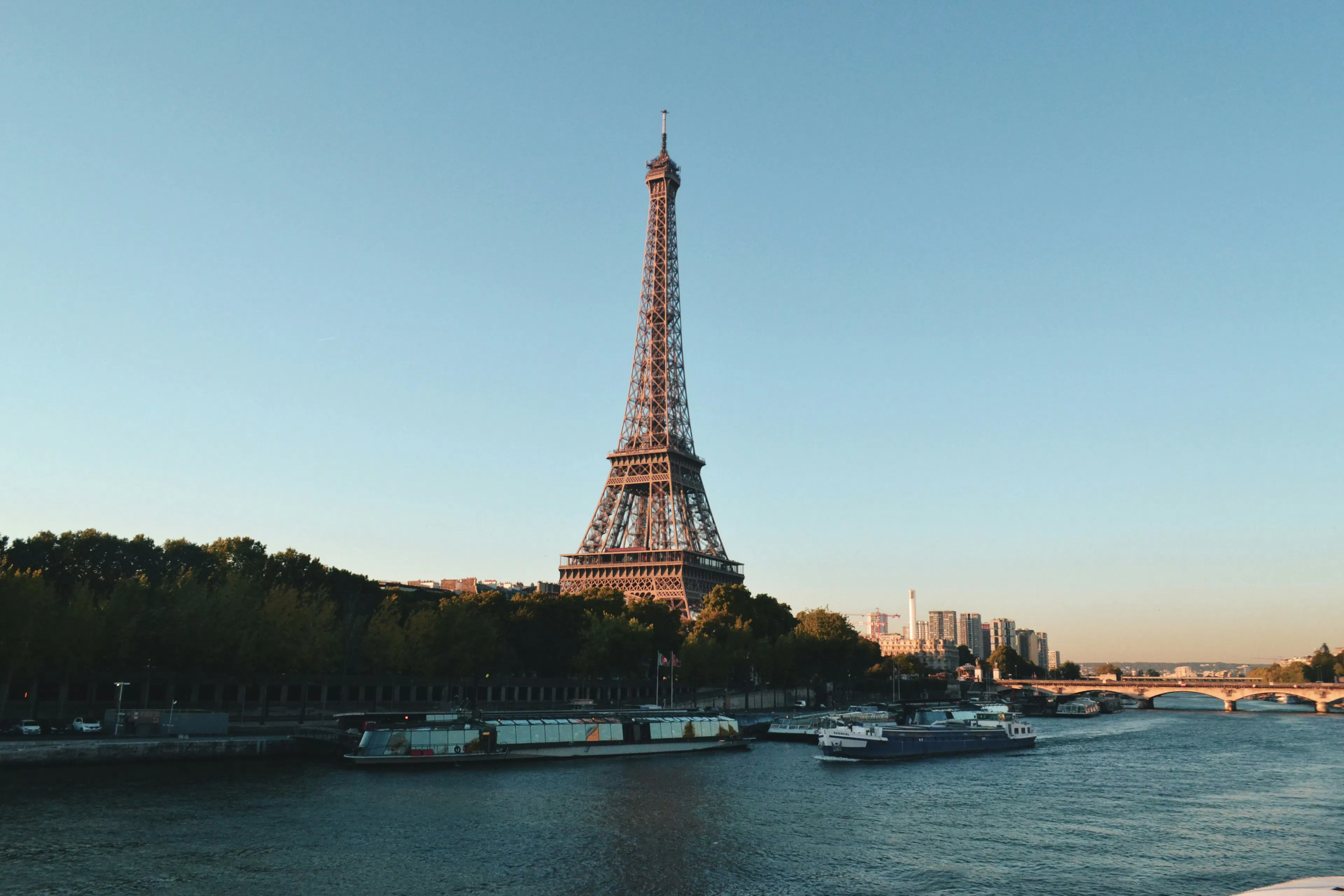 Eiffel Tower rising above the Seine River with boats, trees, and bridges along the waterfront.