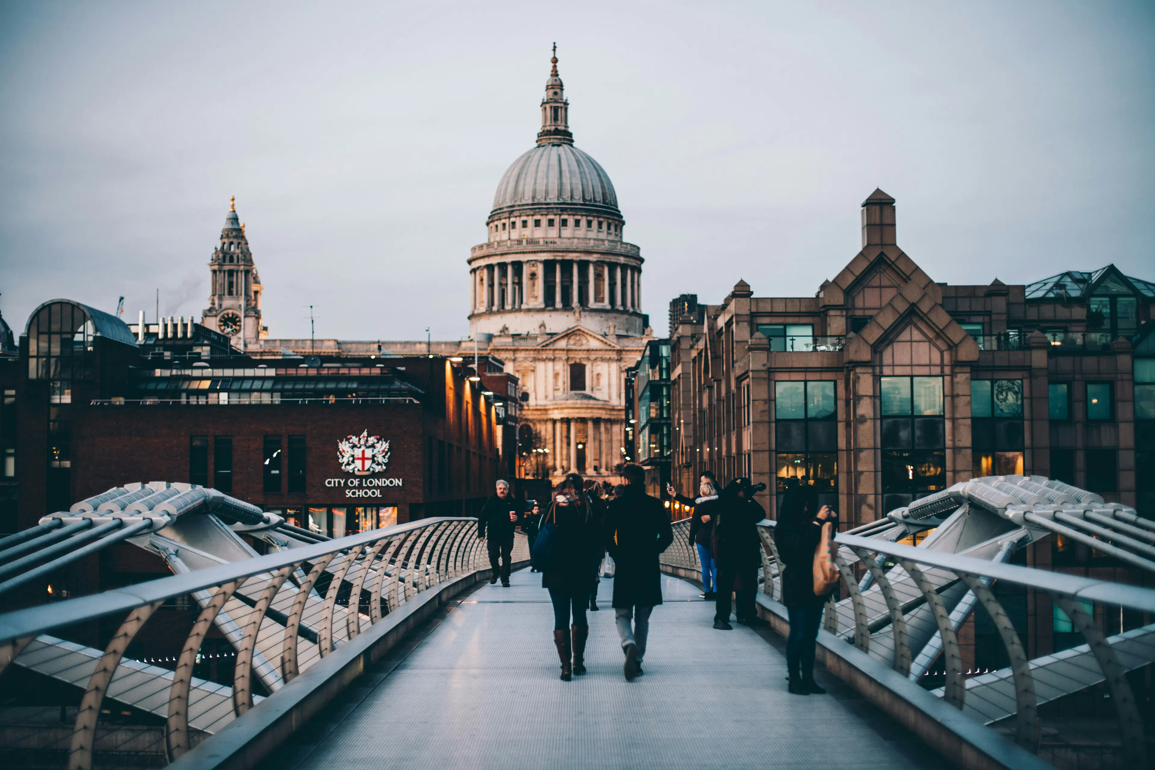Pedestrians crossing Millennium Bridge with St. Paul's Cathedral rising in the background among London buildings.