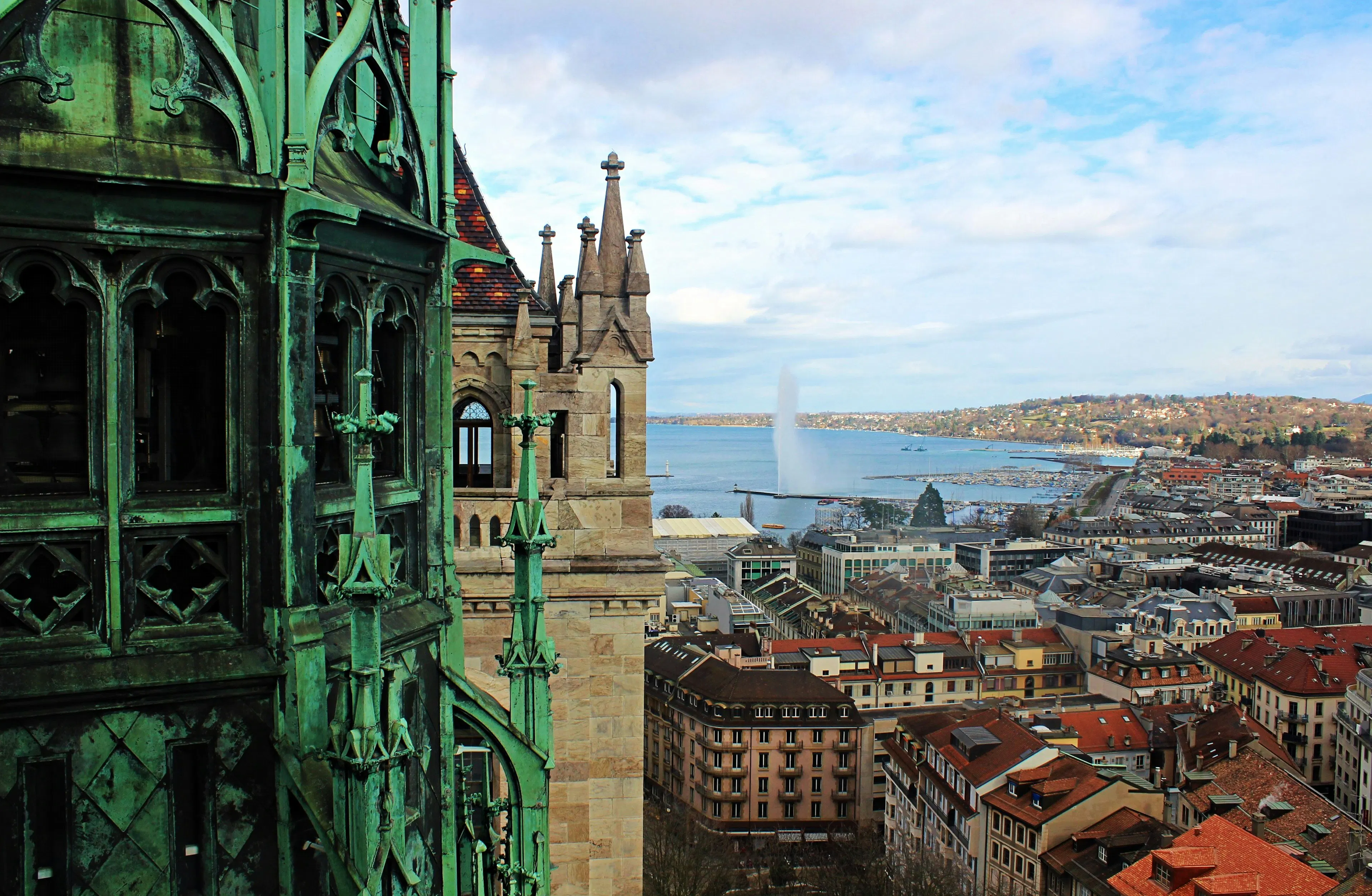 View of Geneva, Switzerland, from a historic stone and copper tower overlooking the city, Lake Geneva, and the Jet d’Eau fountain in the distance.