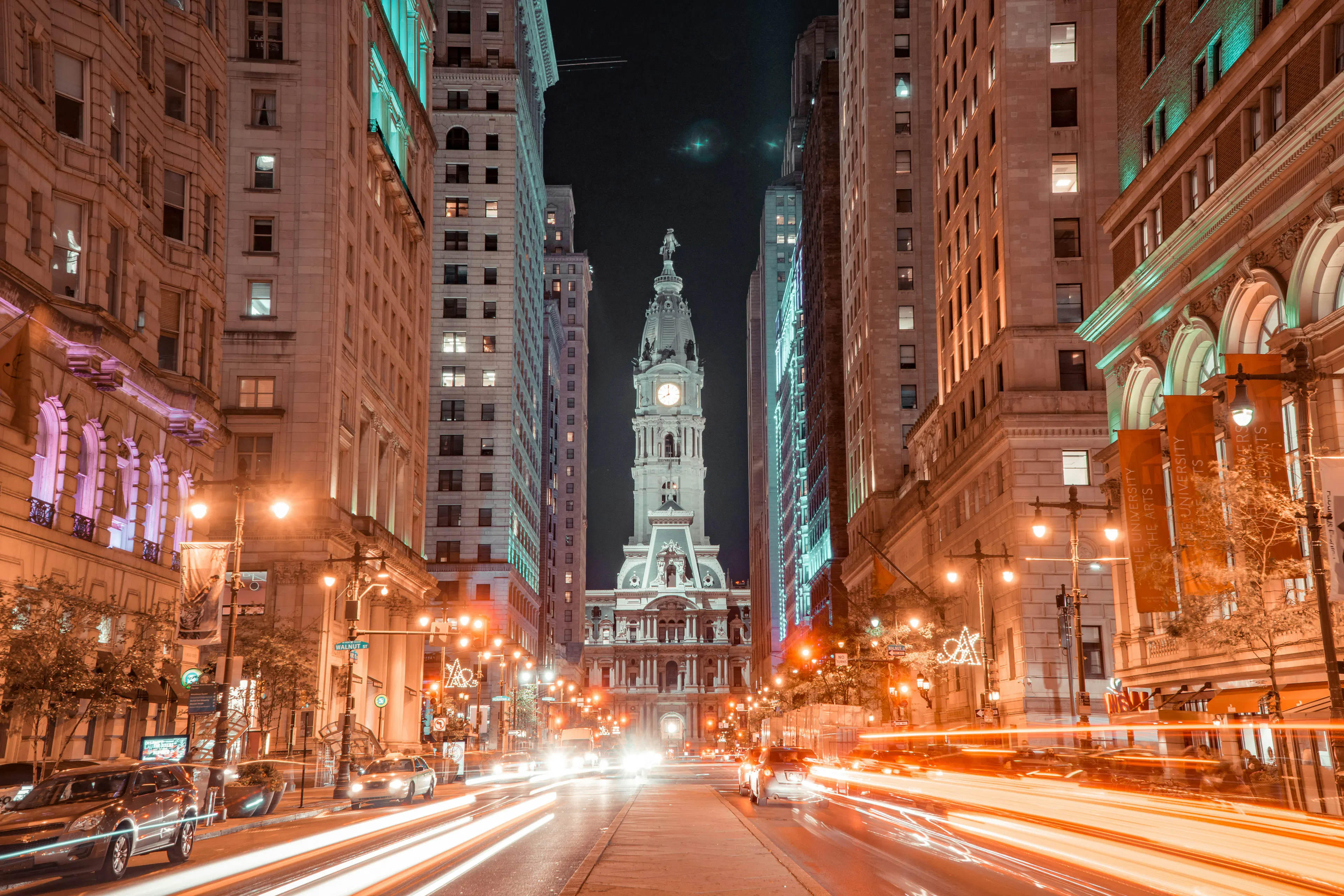 Philadelphia City Hall illuminated at night, framed by downtown buildings and streaks of traffic lights along a city street.
