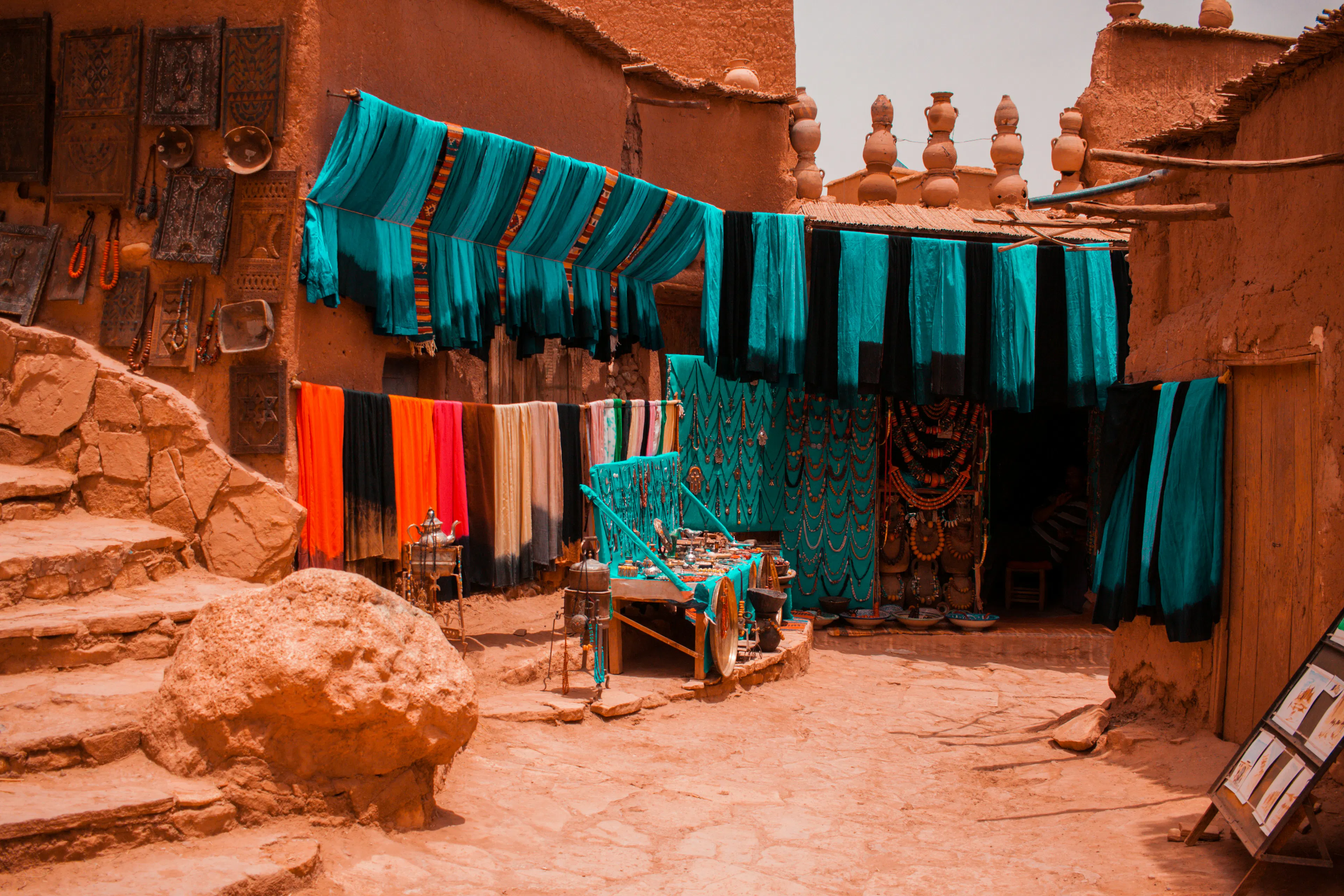 Colorful textiles and handcrafted goods displayed outside a traditional market stall in Marrakech, Morocco, surrounded by earthen buildings and stone walkways.