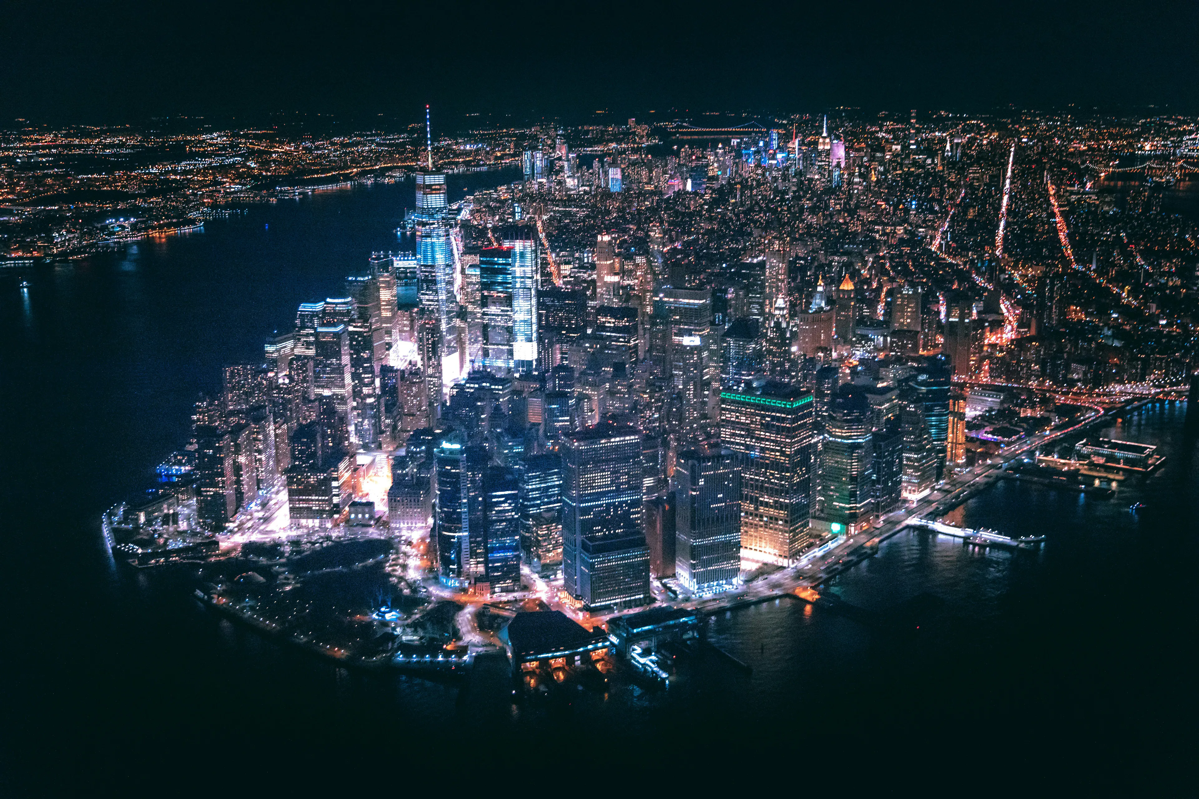 Aerial view of Lower Manhattan at night with illuminated skyscrapers, One World Trade Center, and surrounding waterfront.
