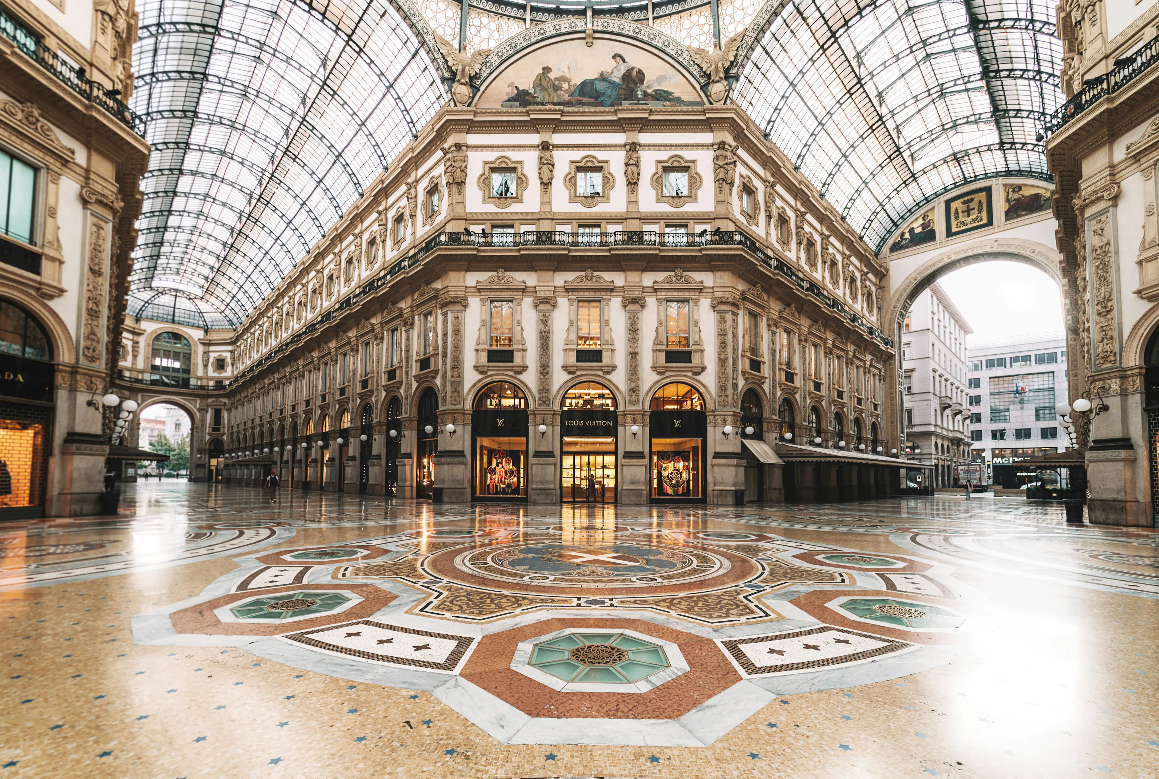 Interior of Galleria Vittorio Emanuele II in Milan, featuring ornate architecture, a glass-vaulted ceiling, and decorative mosaic flooring lined with storefronts.